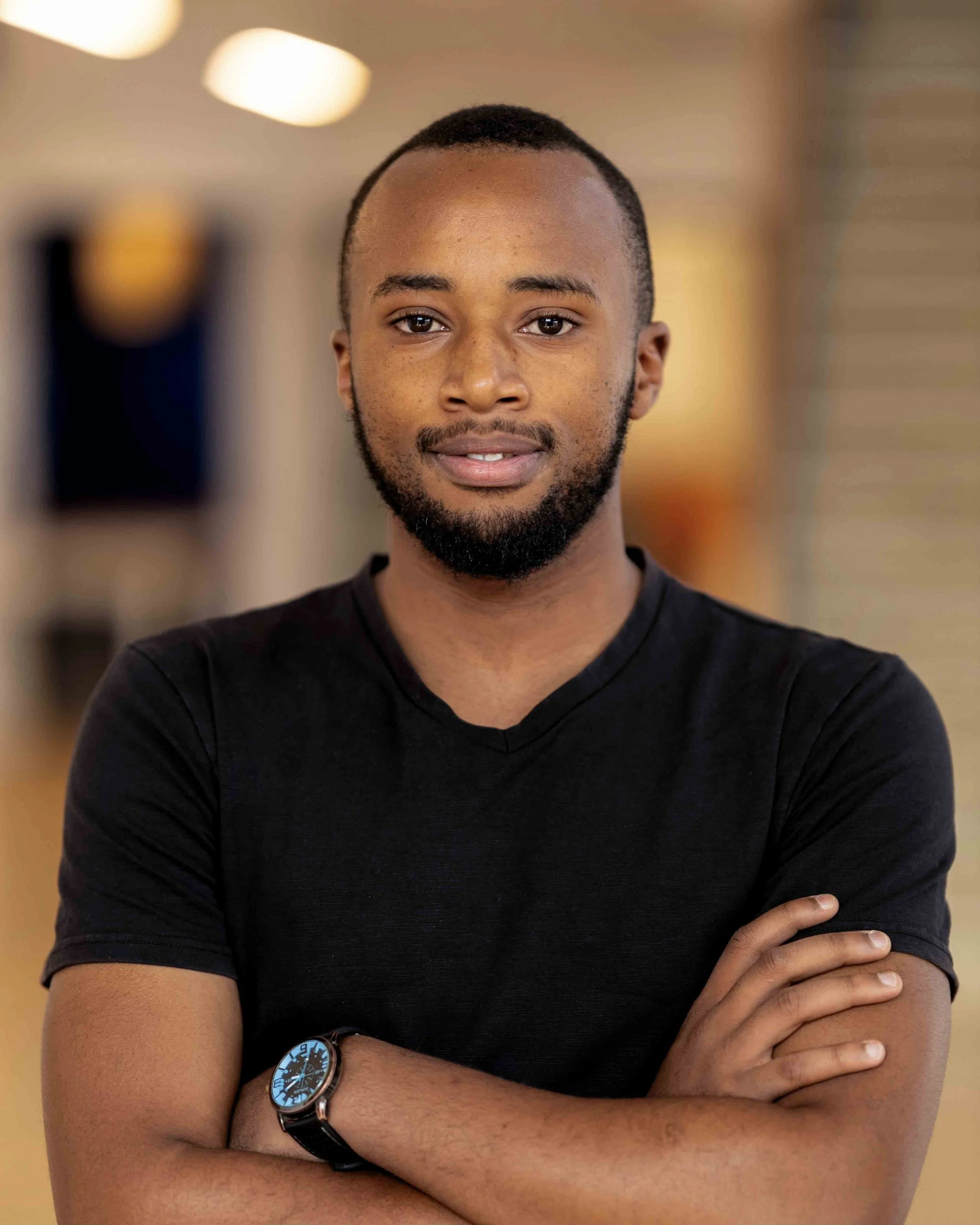 A young man with a short beard and mustache, wearing a black T-shirt and a wristwatch, standing in an indoor setting with blurred background.