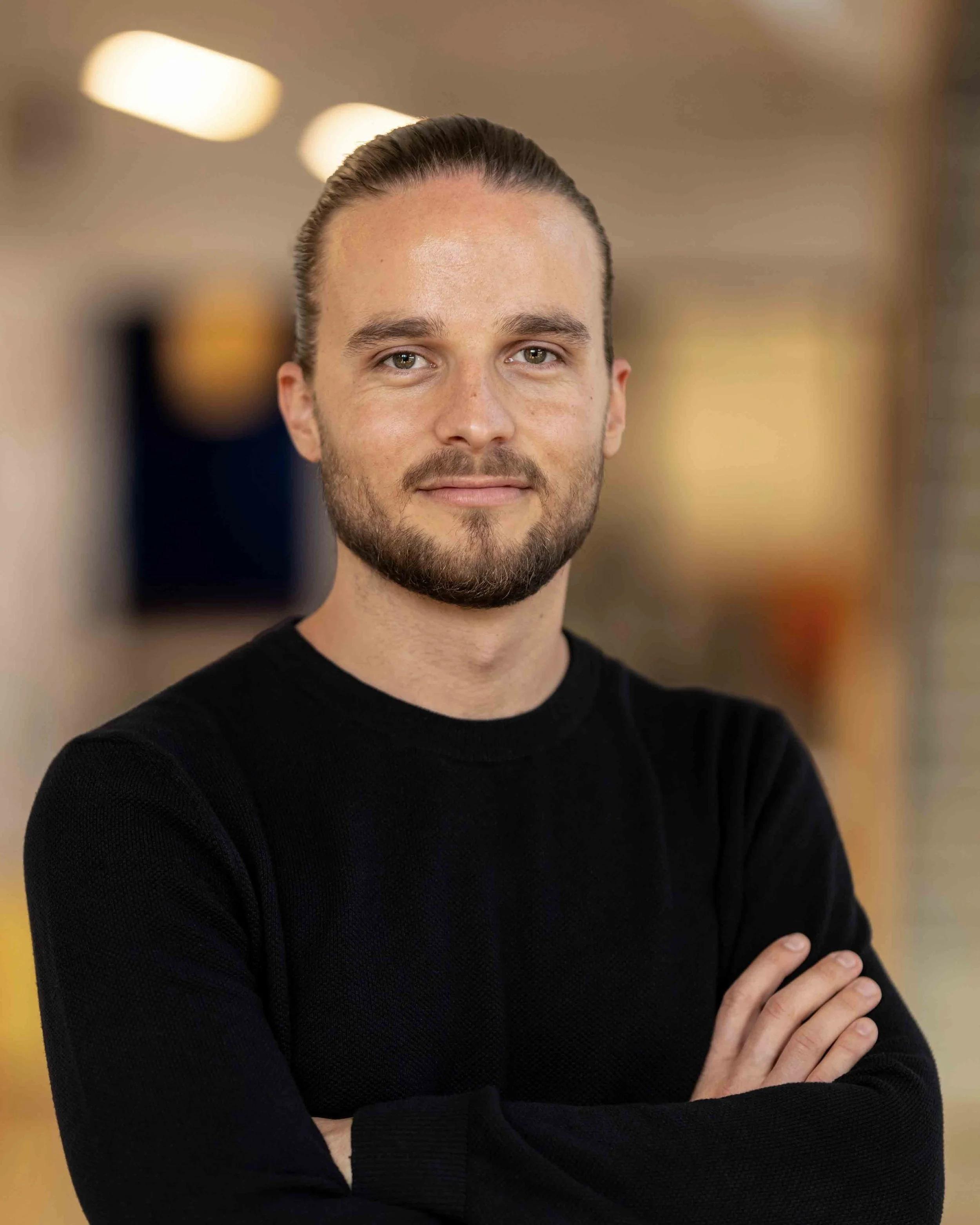A man with a beard and slicked back hair wearing a black sweater, standing with crossed arms in an indoor setting.