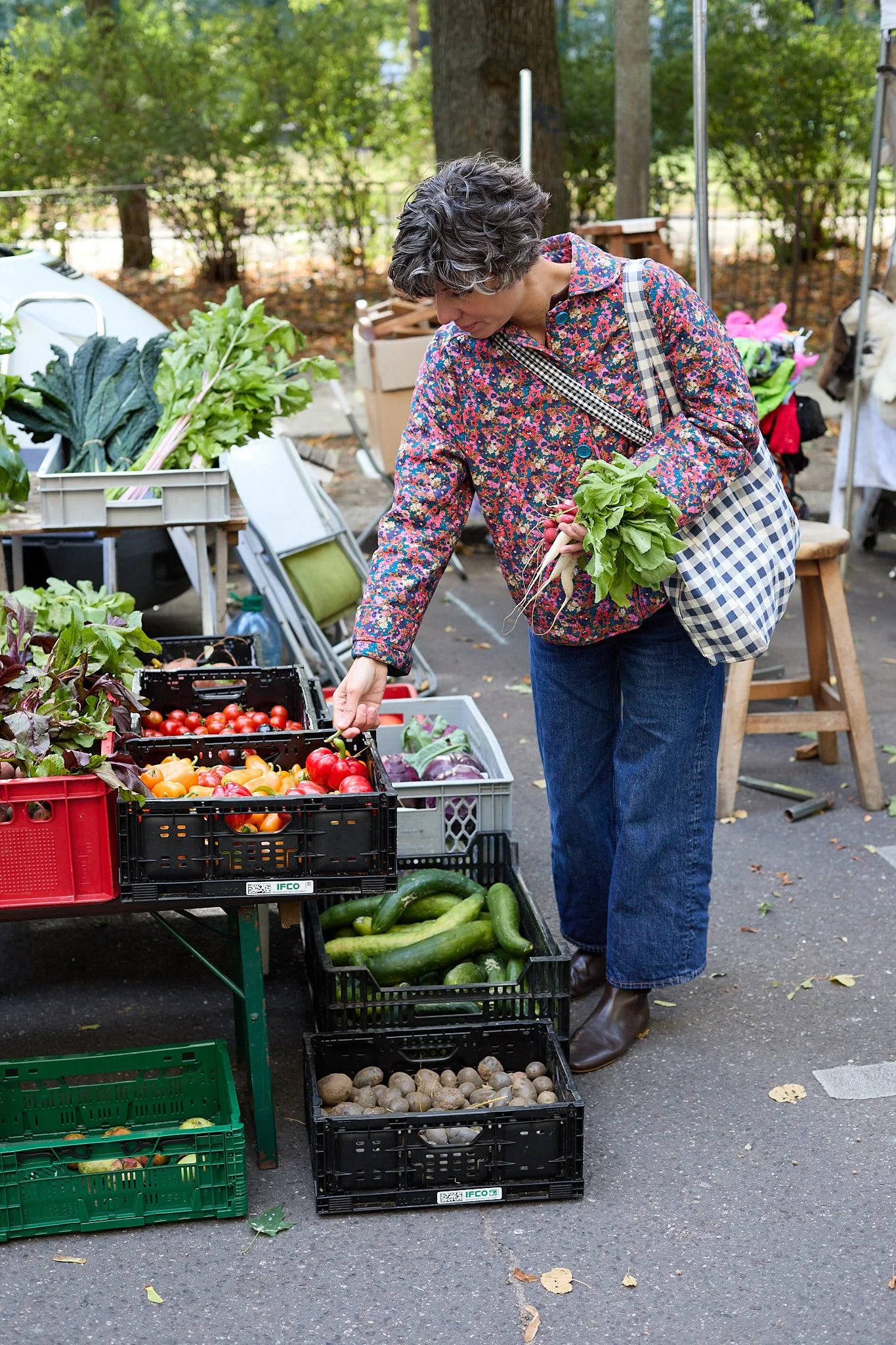 Eine Frau beim Einkauf auf einem Wochenmarkt, sie steht vor mehreren Kisten mit frischem Gemüse wie Tomaten, Salat, Radieschen, Gurken, Kartoffeln und anderem, und füllt ihre Einkaufstasche.