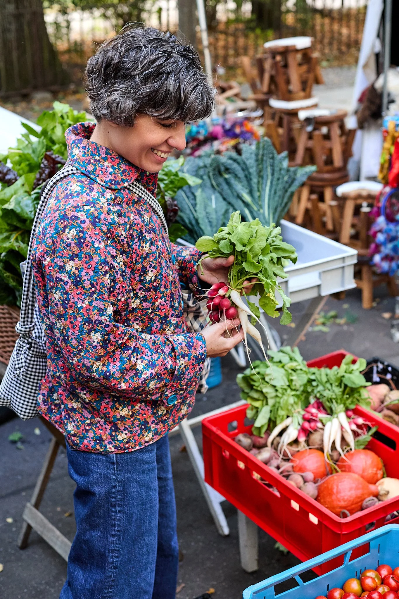 Food Fotografie für Markt Kochbuch: Junge Frau beim Einkauf auf einem Markt, hält einen Bund Radieschen in der Hand, im Hintergrund sind Gemüse und Obst in Körben und Kisten. Sie trägt eine bunte, floral gemusterte Jacke und hat kurze, wellige Haare.