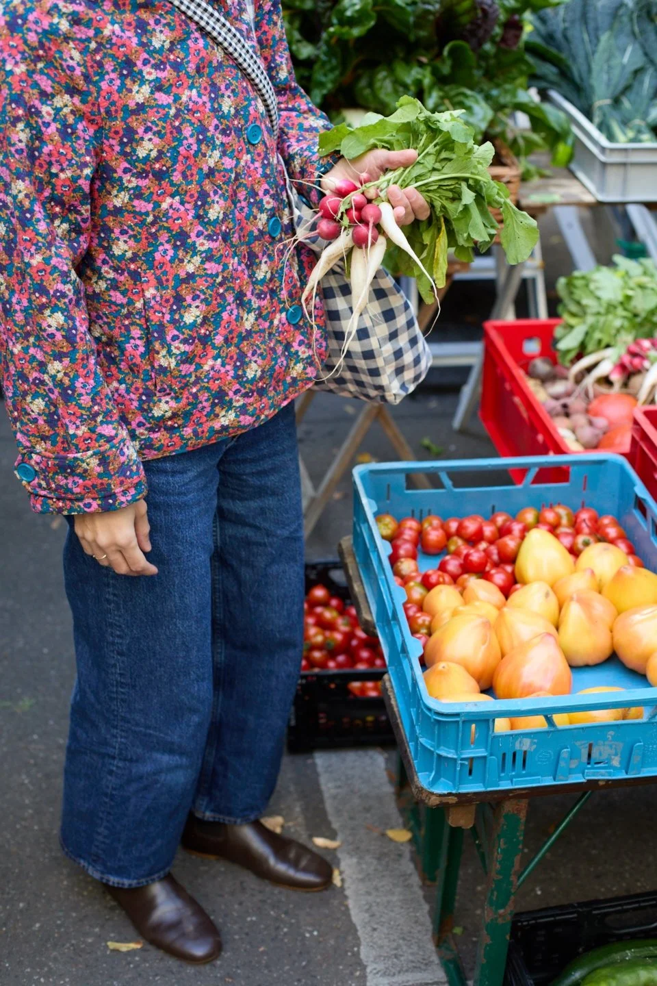 Eine Person hält frische Radieschen und Weiße Rüben mit Blättern in der Hand, an einem Marktstand voller frischer Tomaten, auf einem Bauernmarkt.