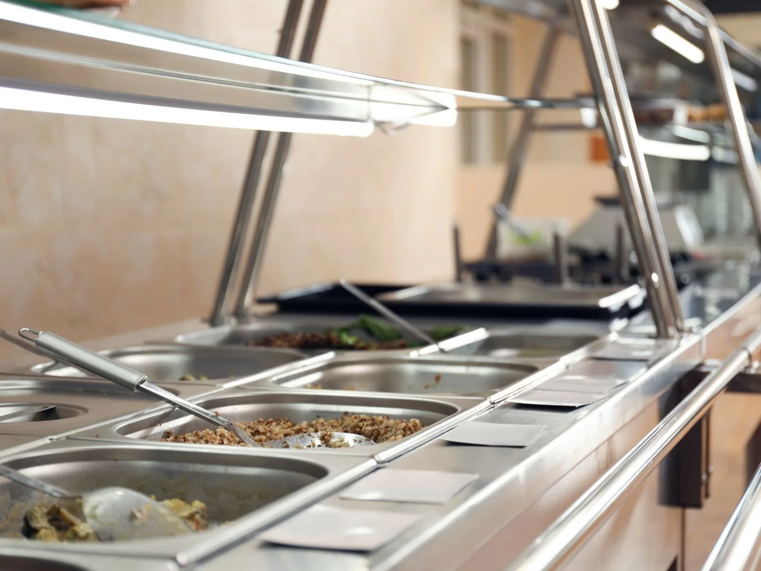 Une sélection de plats chauds et froids dans un service de buffet, avec plusieurs plats en inox et des cuillères, dans une ambiance de cafétéria ou restaurant.