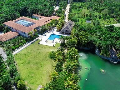 Aerial view of a resort with a large building, swimming pool, gardens, and a water body surrounded by lush greenery.