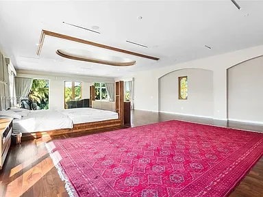 A spacious bedroom with a large window and a white bed, adjacent to a vibrant pink area rug on wooden flooring, with white walls and ceiling.