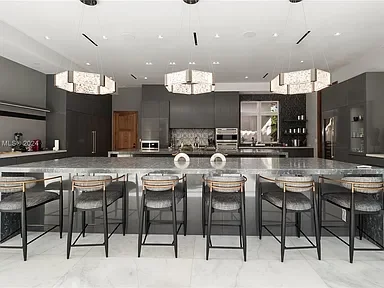 Modern kitchen with gray cabinets, white countertops, and a marble island with seven barstools. Three rectangular chandeliers hang above the island.