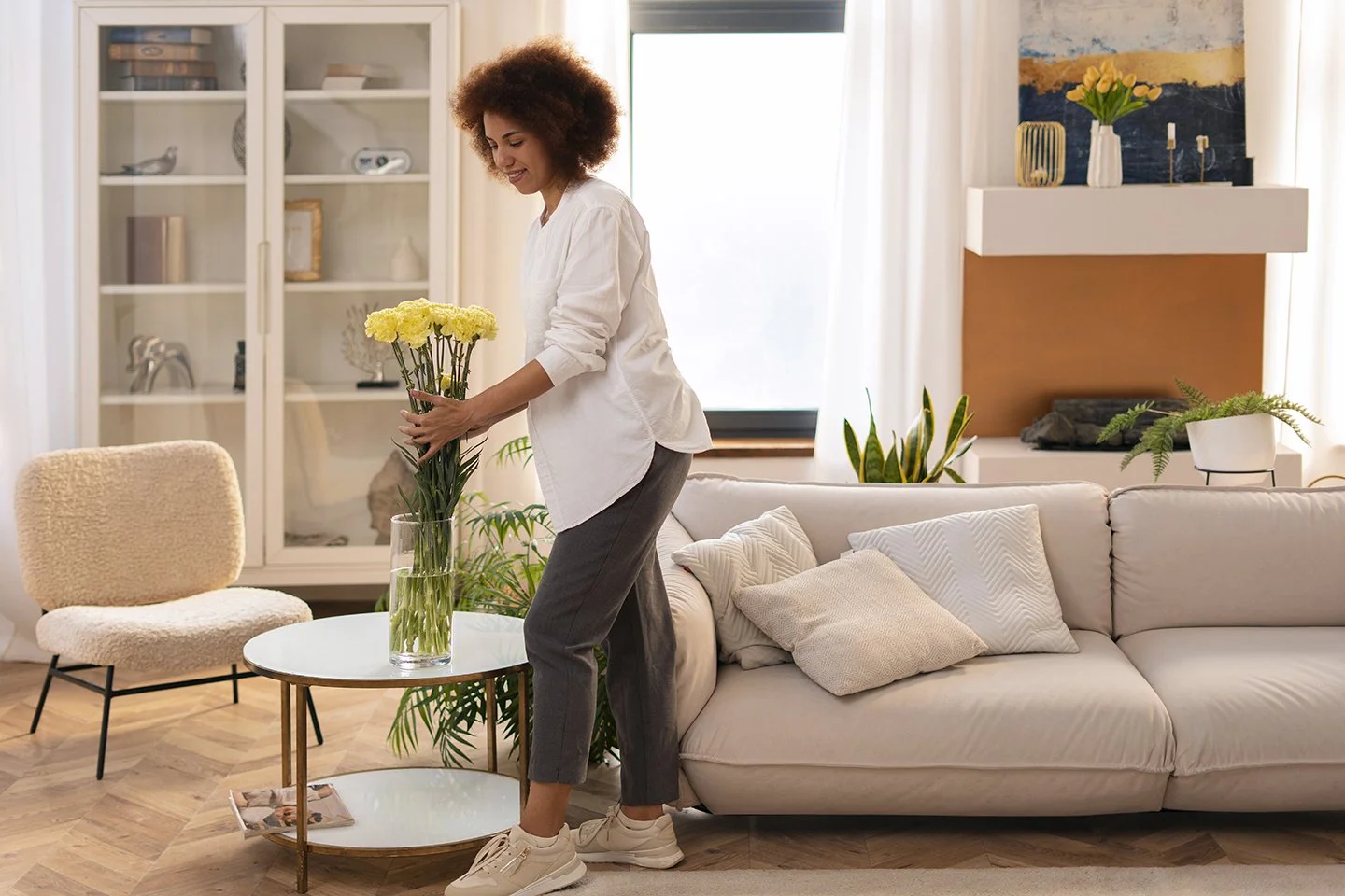 A woman arranging yellow flowers in a living room with a white sofa, plants, and decor.