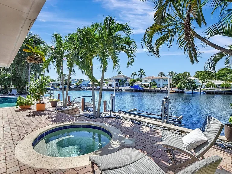 View of a backyard patio with two chairs and a small hot tub, overlooking a canal with boats and waterfront houses, under a sunny sky with some clouds.