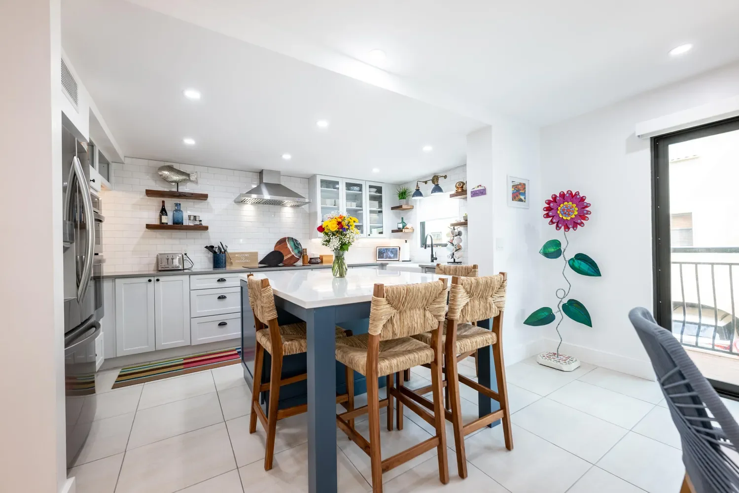 Modern white kitchen with island, wooden bar stools, and colorful flower bouquet on the counter. Open shelves, stainless steel appliances, and a patio door with balcony visible.