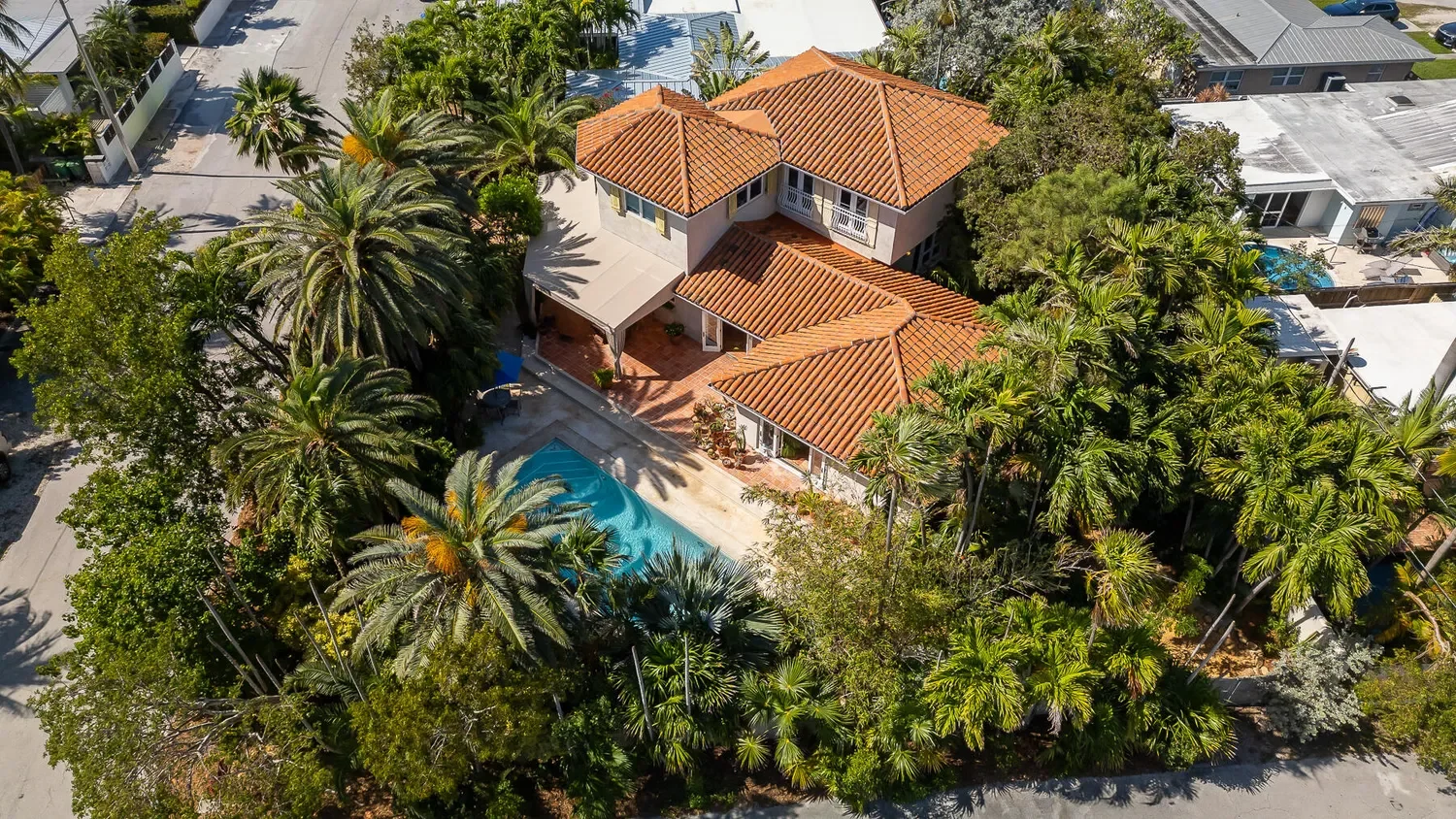 Aerial view of a house with a red-tile roof, surrounded by lush green trees and palm trees, featuring a swimming pool in the backyard.