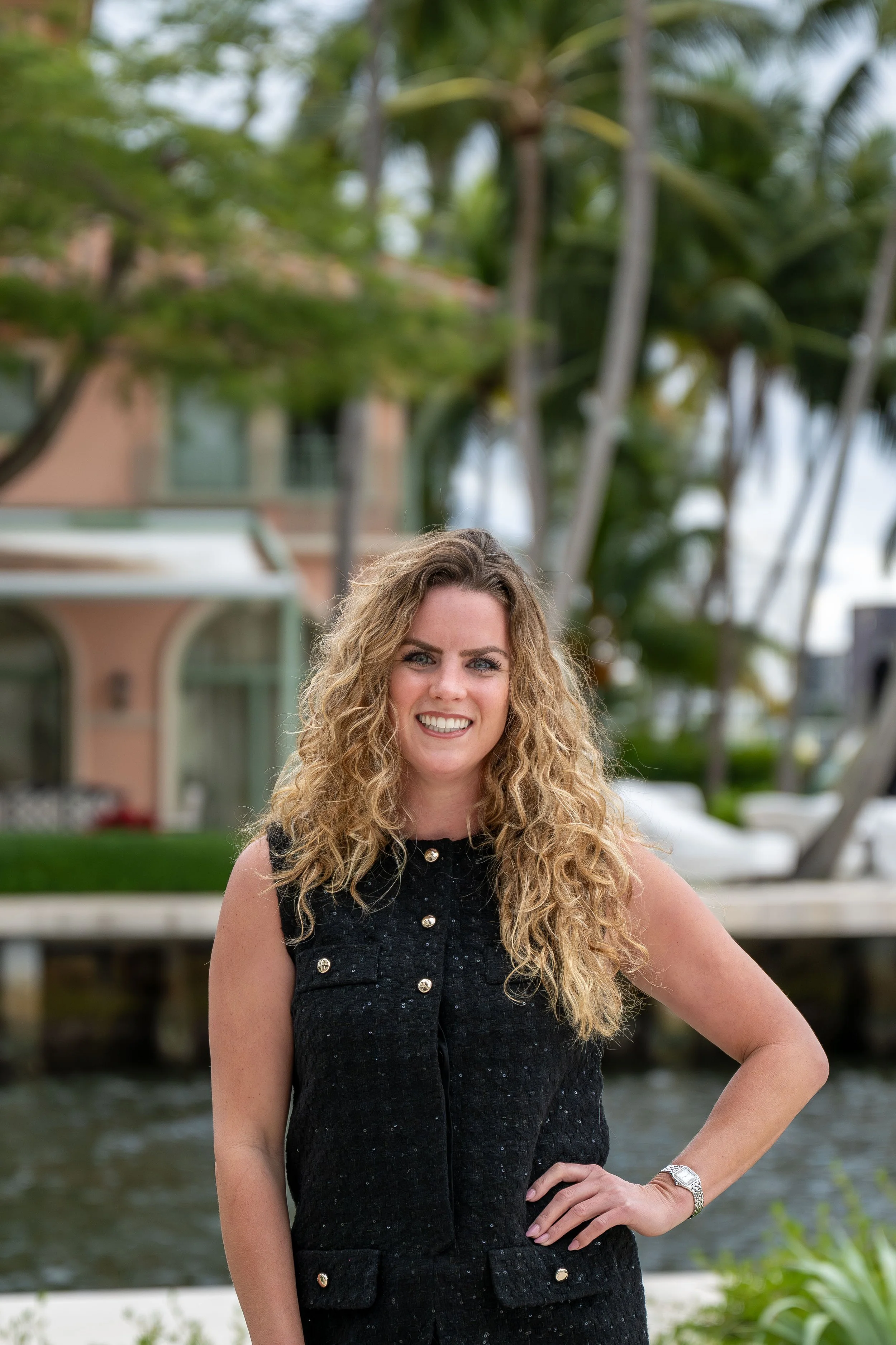 A woman with curly blonde hair standing outdoors near water, smiling, with palm trees and buildings in the background.