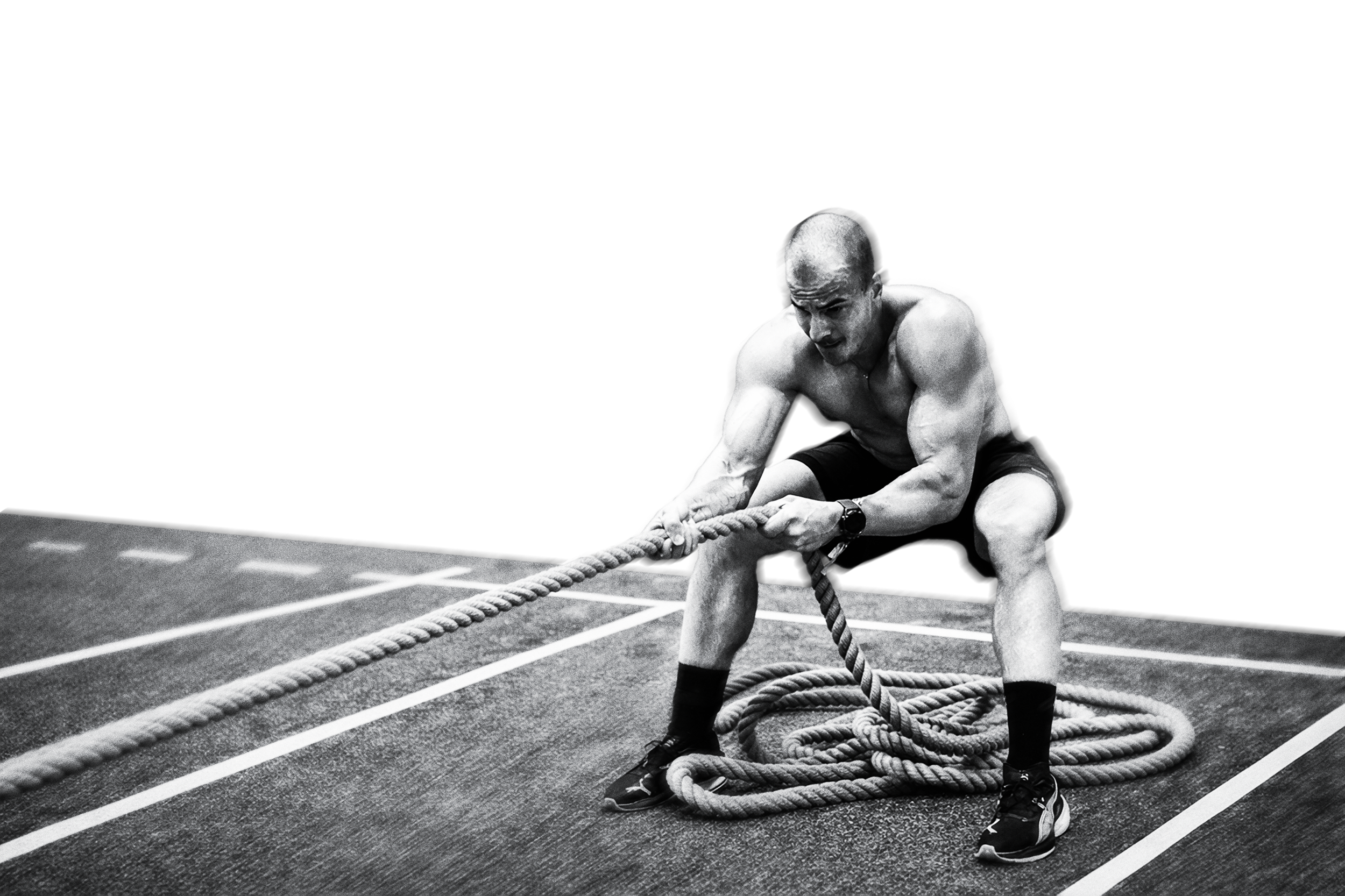 A muscular man, dressed in shorts, sneakers, and a watch, performs a rope exercise on a gym floor marked with white lines, with a serious expression.