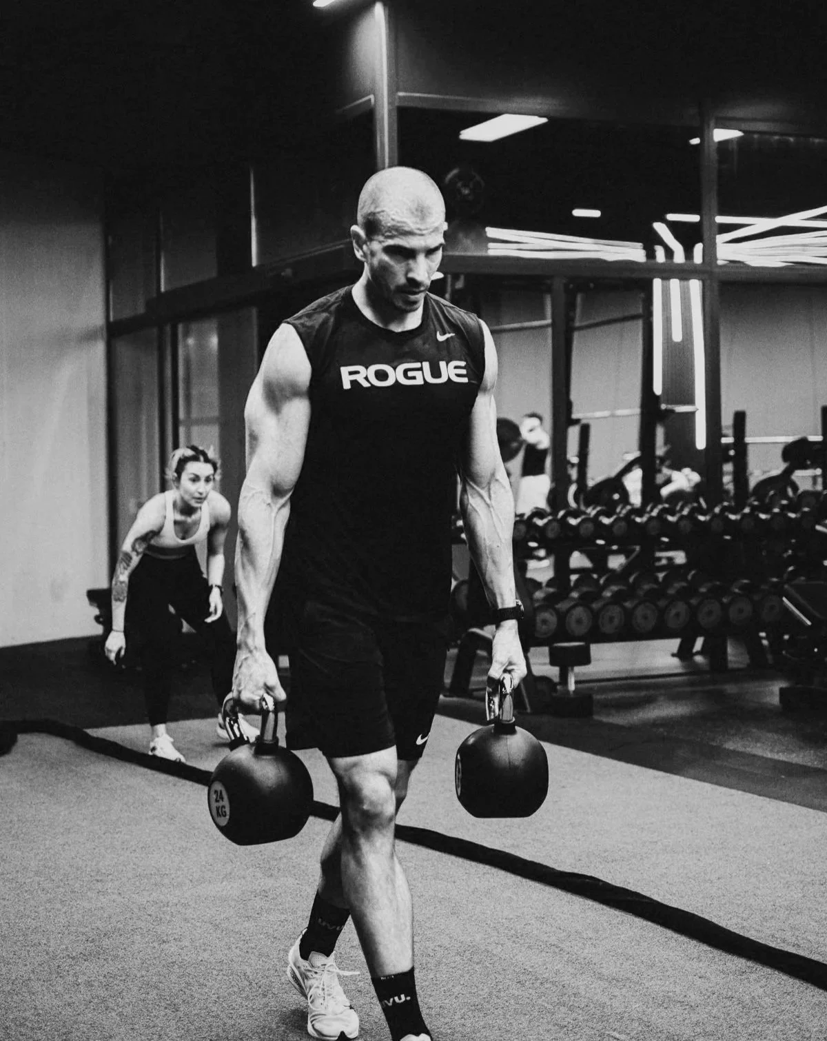 A man in a black athletic shirt and shorts carrying two kettlebells at a gym. A woman is in the background, also exercising.