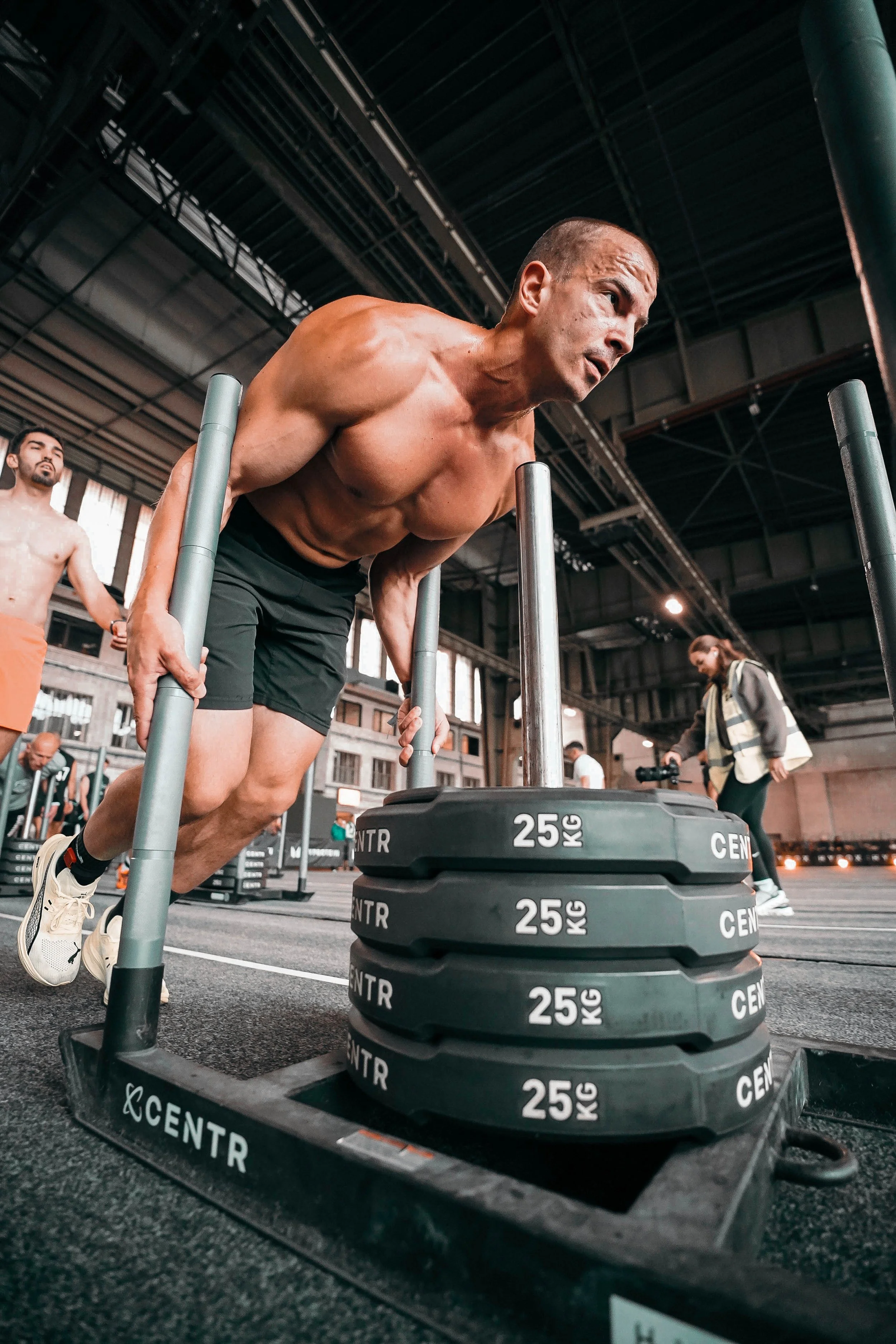 Man performing push-ups with a weighted sled in a gym, surrounded by people and gym equipment.