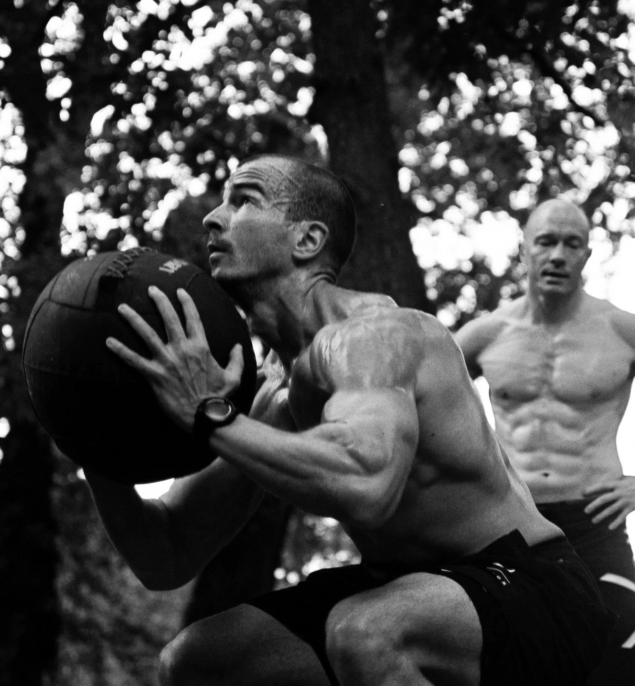 A shirtless man with a muscular build holding a medicine ball close to his chest outdoors, with another shirtless man watching in the background.
