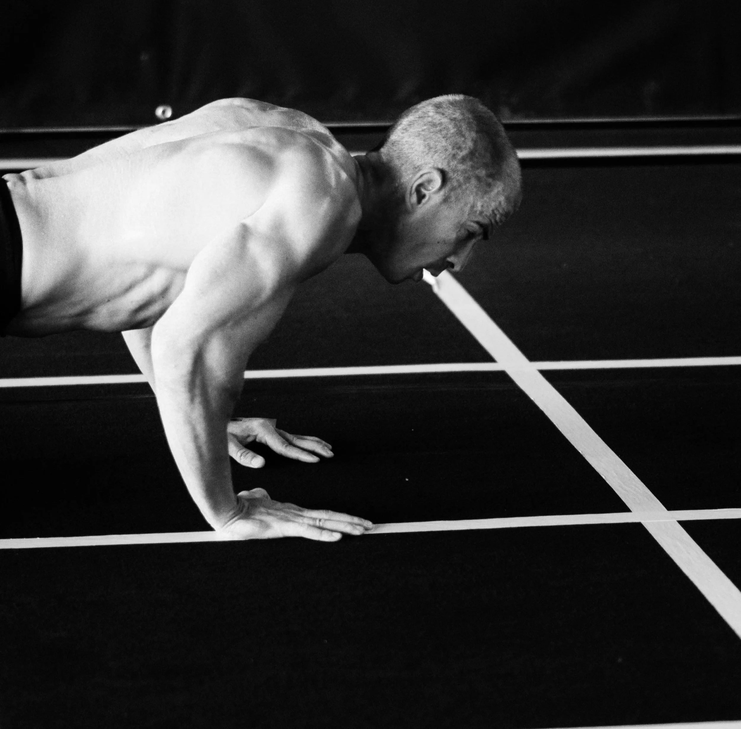 A man with a short haircut and a muscular build doing push-ups on a sports court with white boundary lines, in black and white.