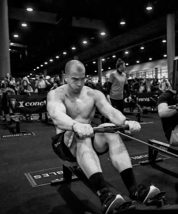 A shirtless man with a shaved head rowing on a rowing machine in a gym or fitness center filled with people and exercise equipment.