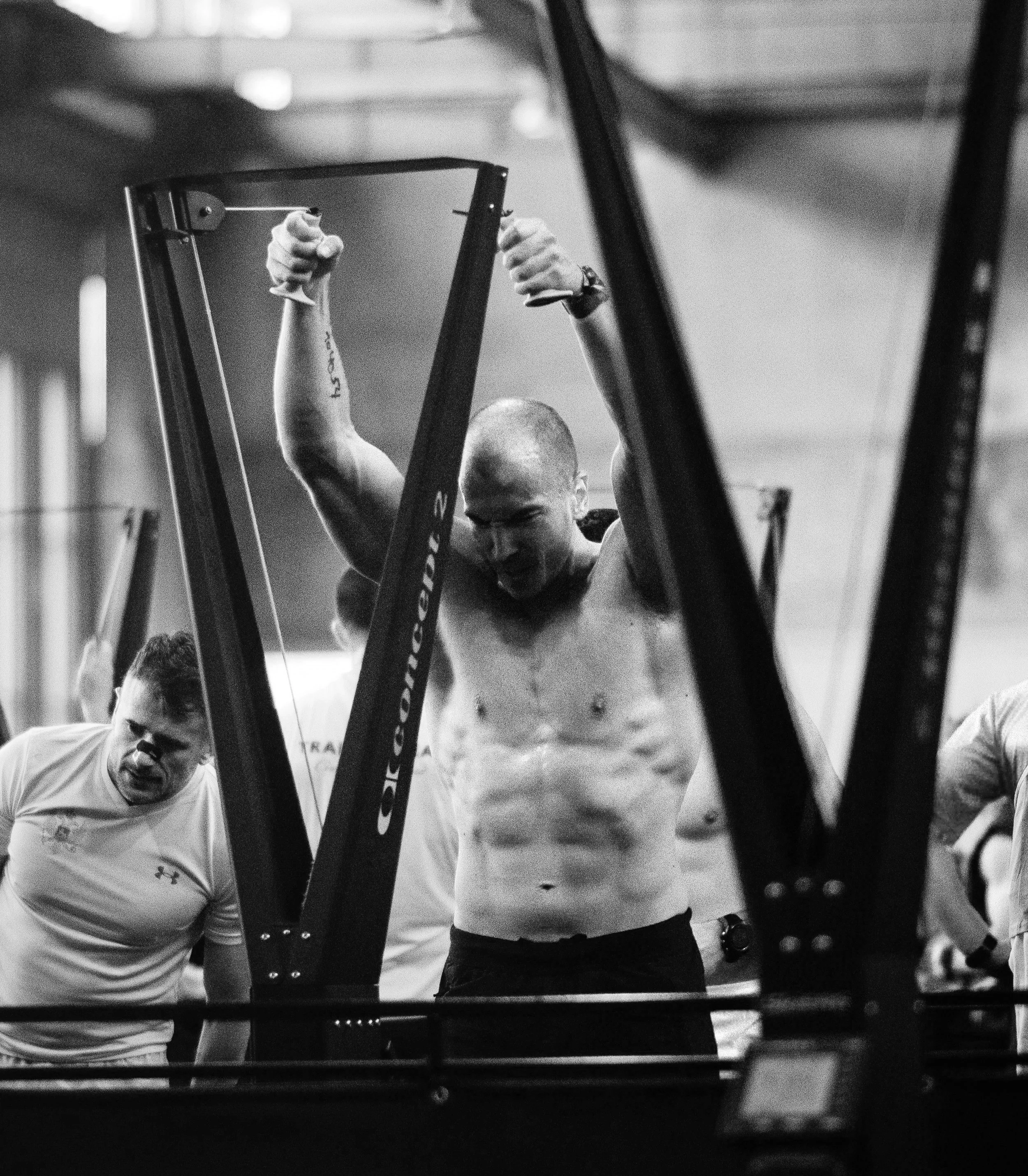A shirtless man with a shaved head exercising on a grappling machine, with his arms raised and gripping the handles, surrounded by other individuals in a gym.