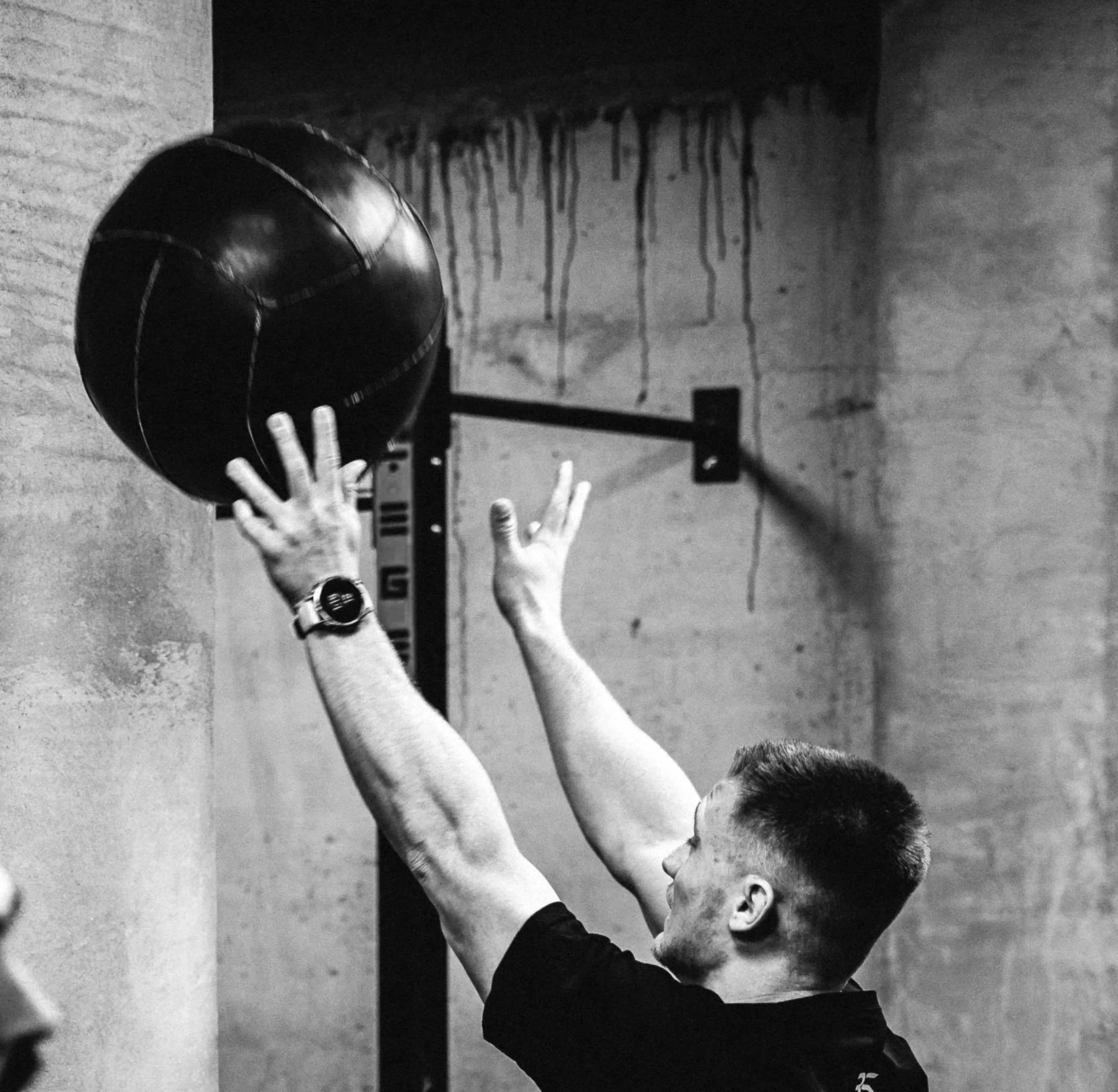 A man is performing a wall ball exercise, tossing a medicine ball against a wall in a gym.