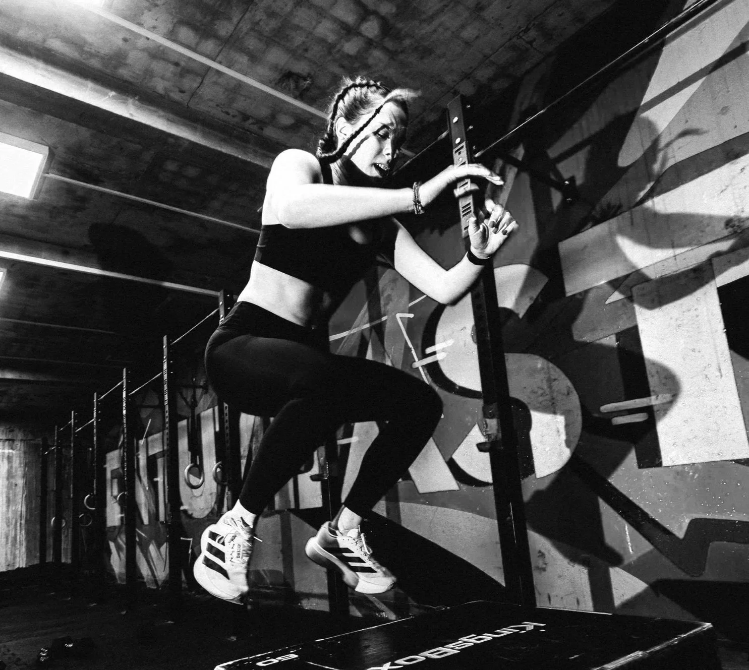 A woman in workout clothes, including leggings, a sports bra, and athletic shoes, uses a sledgehammer to break a wooden board at an indoor gym, with gym equipment and graffiti on the wall in the background.