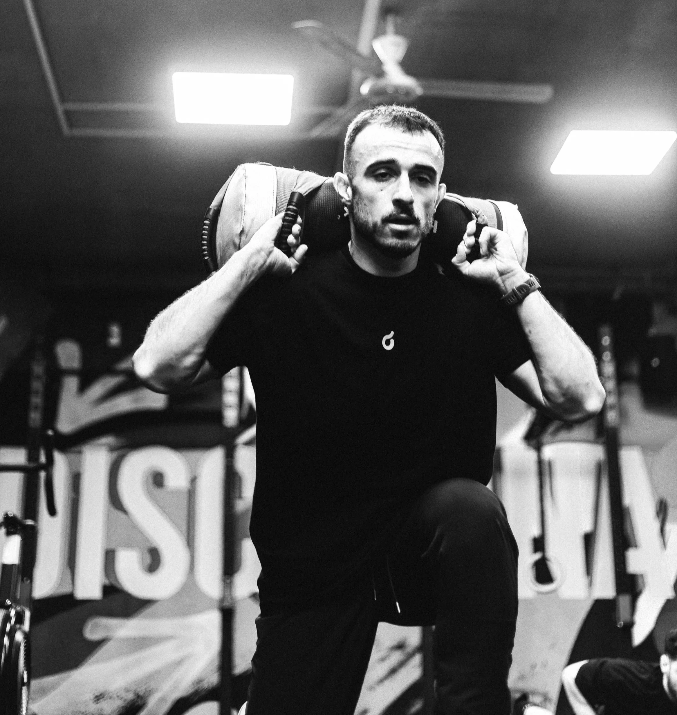 A man carrying a heavy bag on his shoulders inside a gym, with workout equipment and motivational signs in the background.