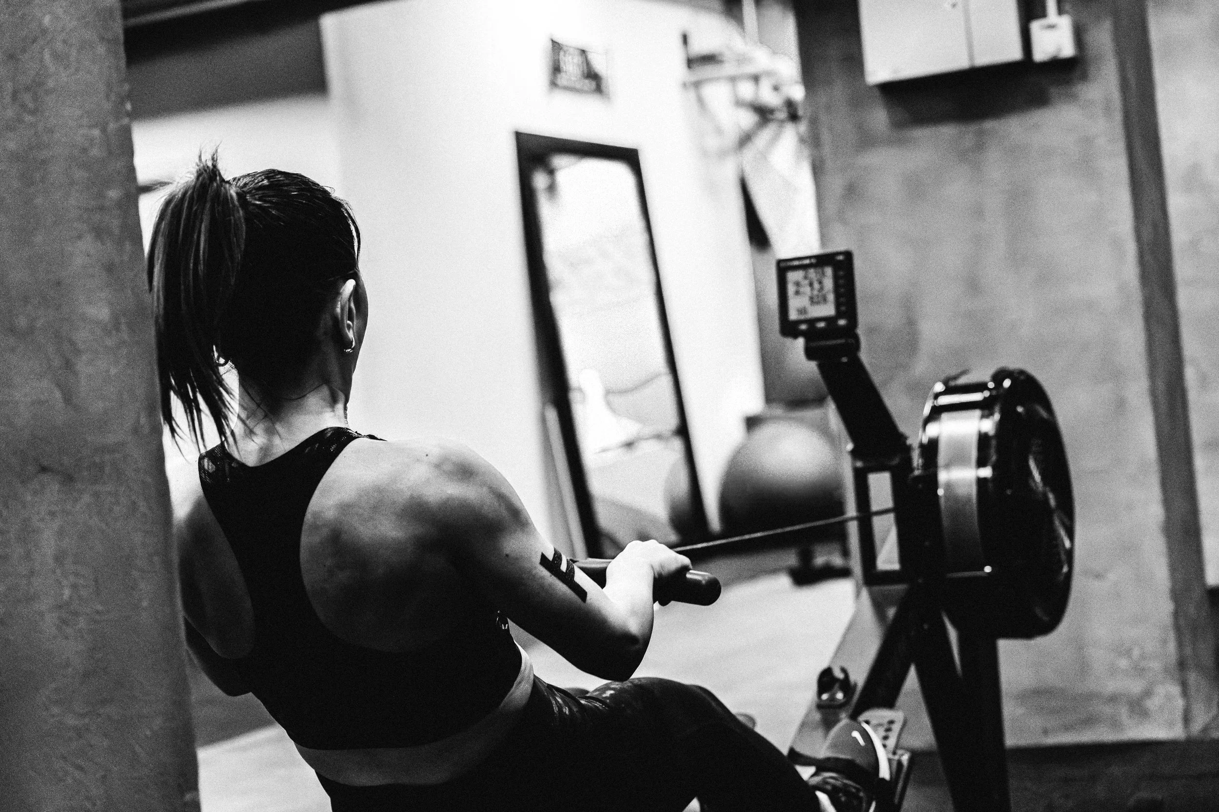 A woman with a ponytail working out on a rowing machine in a gym, with fitness equipment visible in the background.