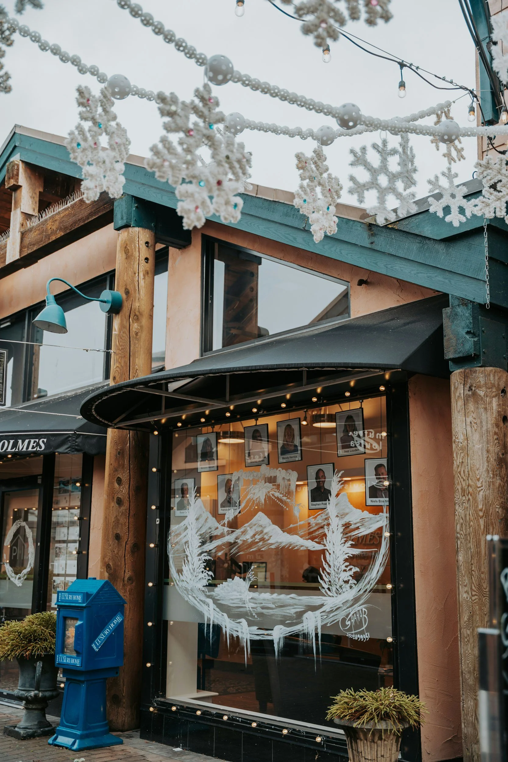 A shop with a large window painted with snow-capped mountains and pine trees, decorated for winter with string lights, snowflake ornaments, and snow-themed decorations on the roof.