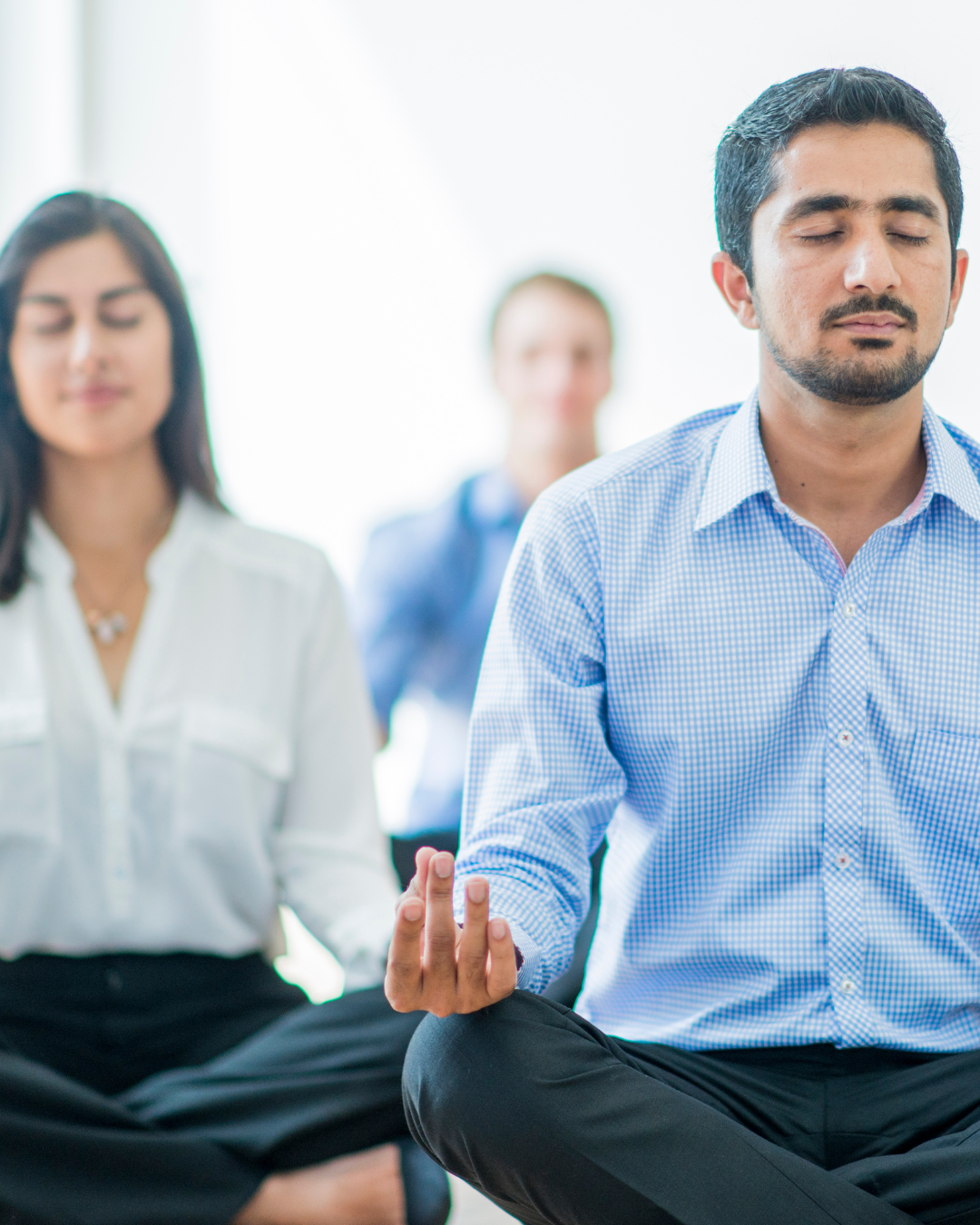 Three professional adults in an office space practicing meditation in a bright, indoor setting, sitting cross-legged with eyes closed, relaxed expressions.