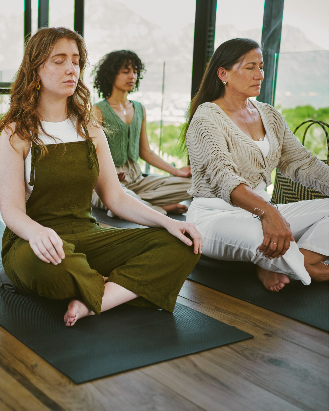 Three women meditating on yoga mats indoors with large windows and a view of greenery outside.