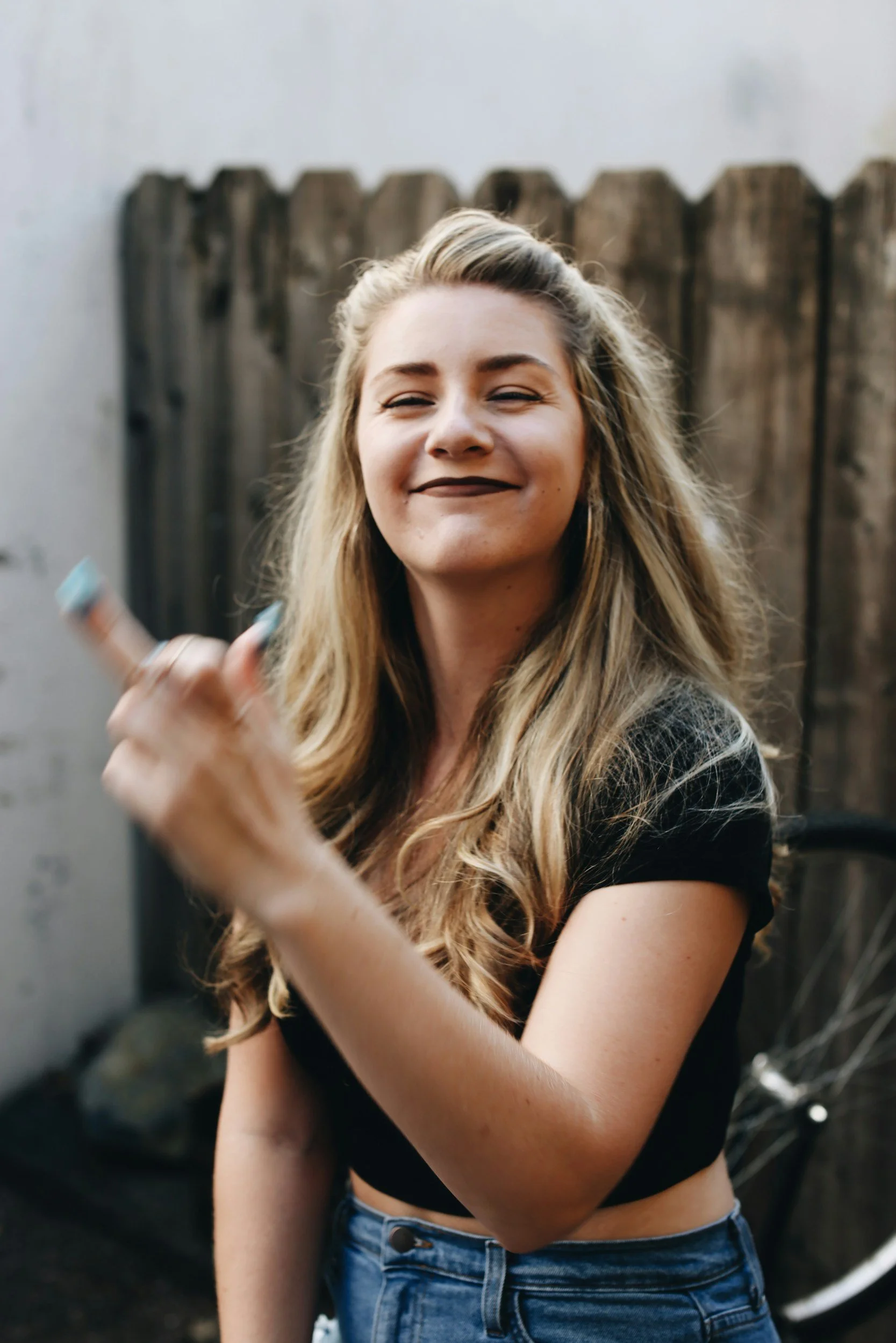 A young woman with long blonde hair smiling and making a hand gesture in front of a wooden fence, with a bicycle in the background.