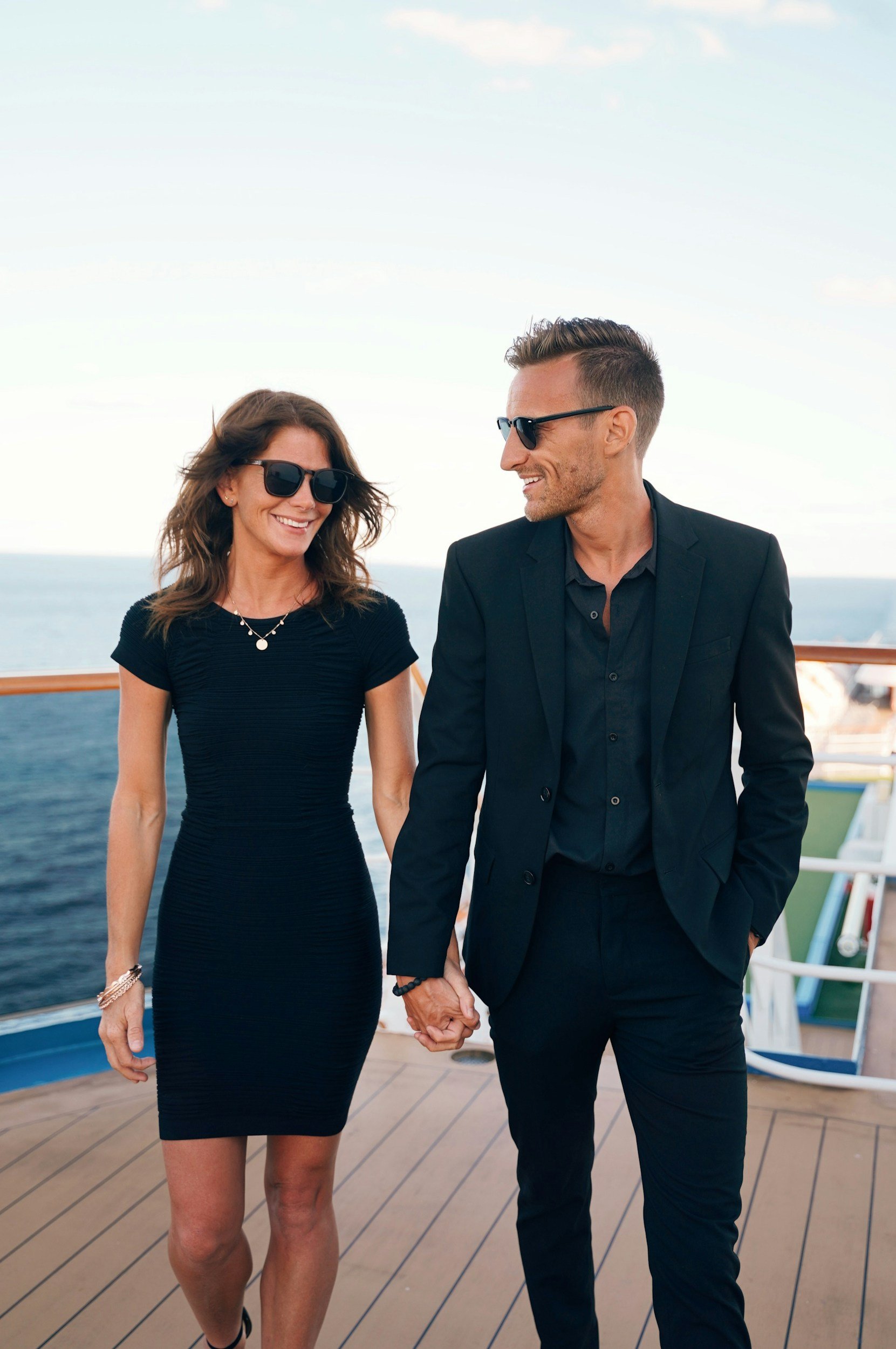 A couple walking hand in hand on the deck of a boat or cruise ship, dressed in elegant black clothing, with the ocean and blue sky in the background.