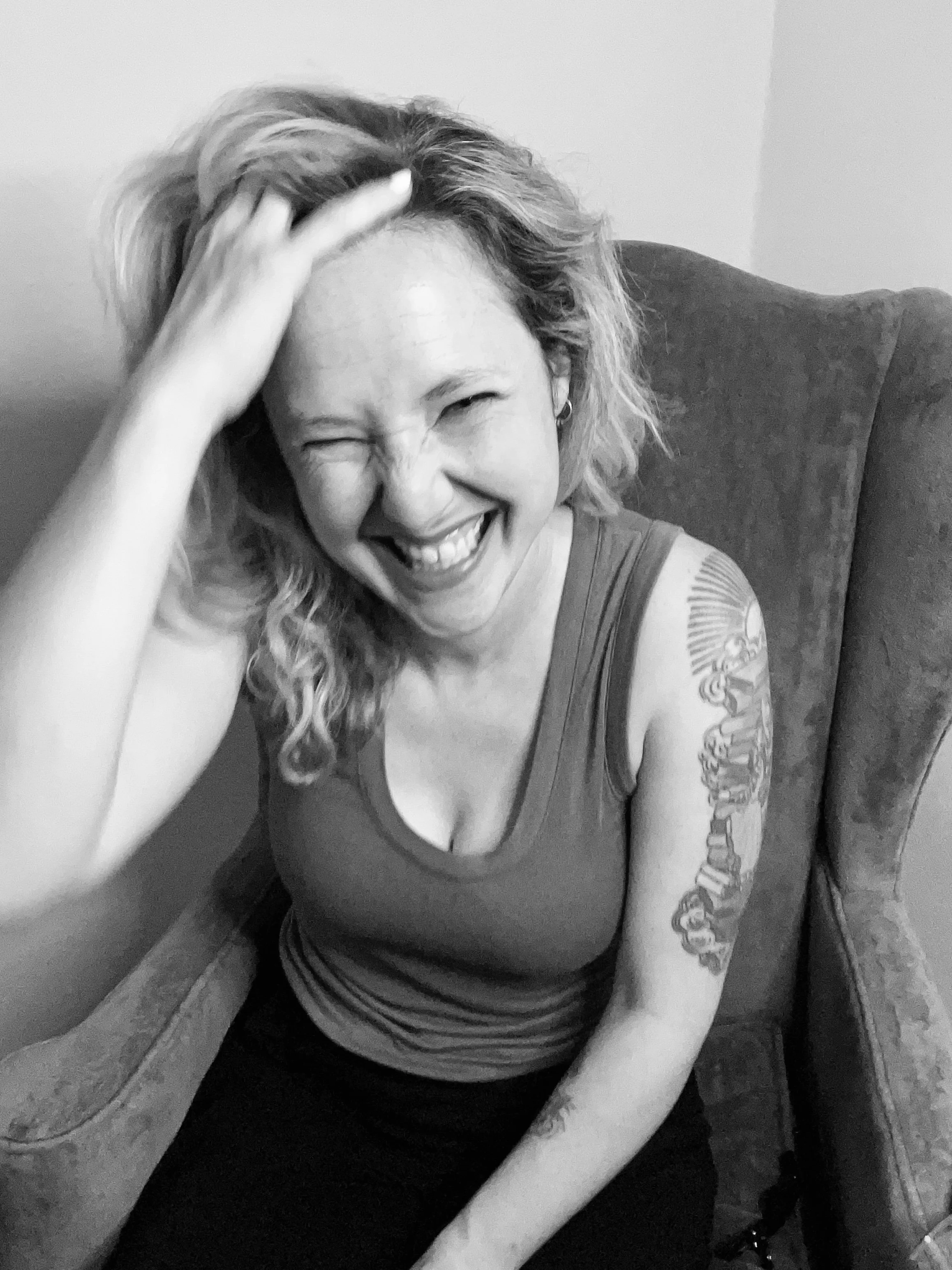 Black and white photo of a woman sitting in a chair, smiling and laughing with one hand on her head, showing tattoos on her upper arm.