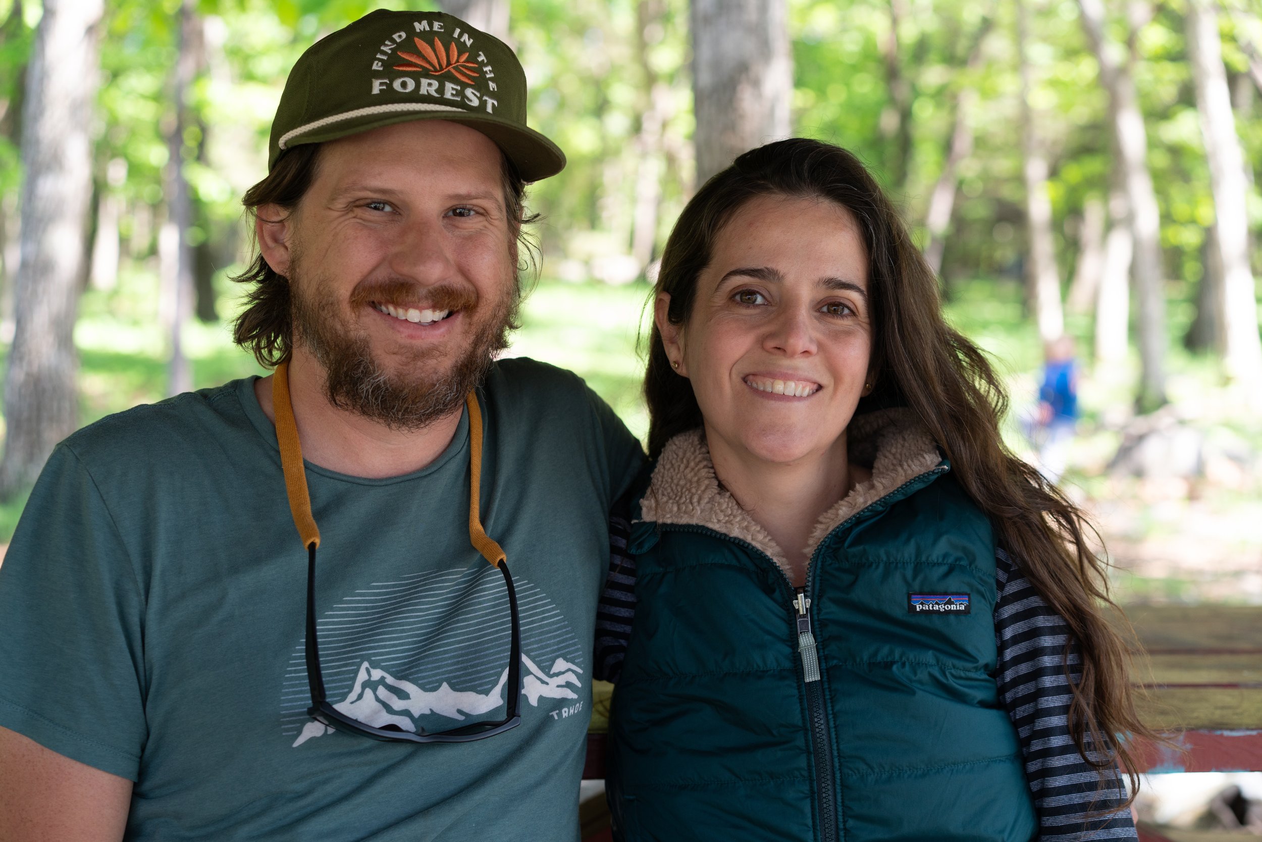 Couple wearing outdoor gear is surrounded by trees and smiles at the camera