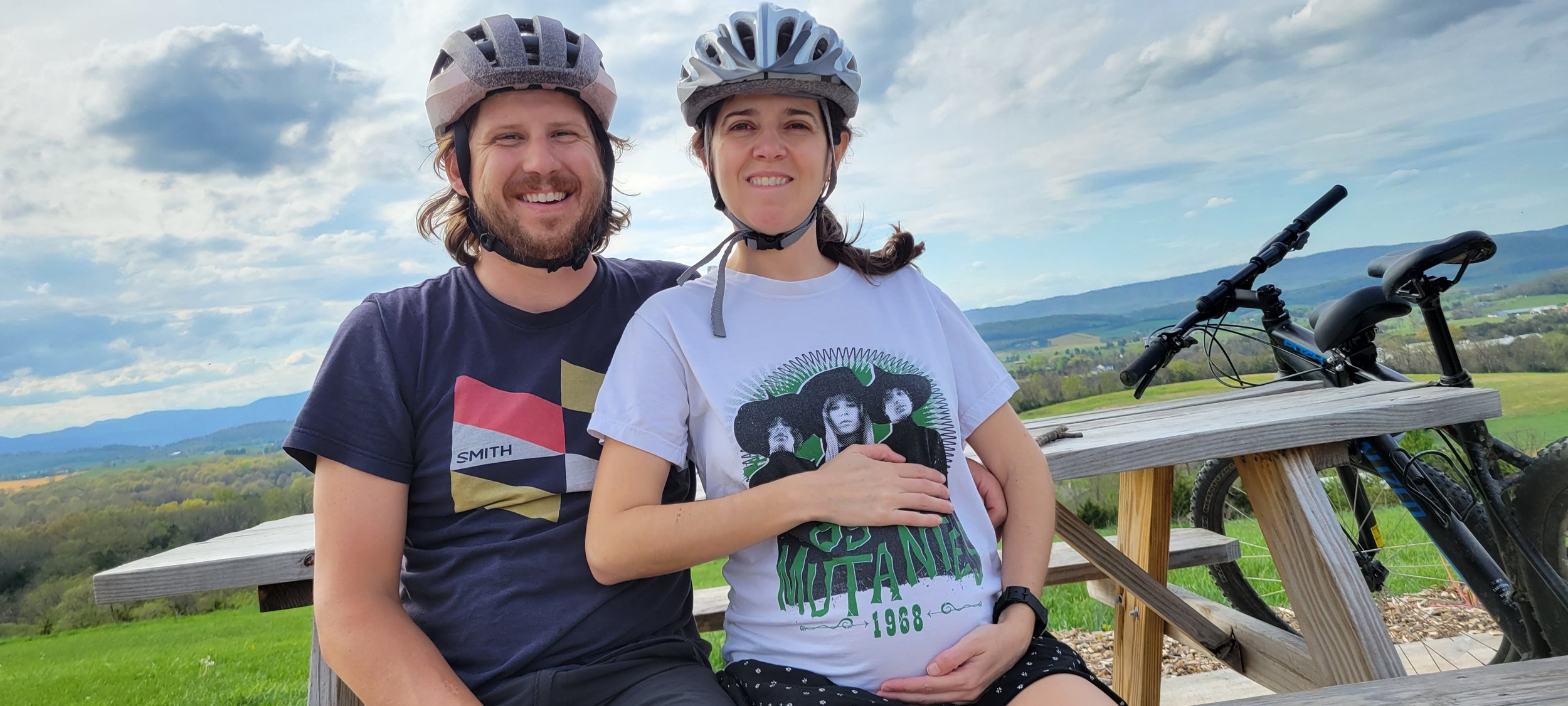 Pregnant woman and man wearing helmets smiling at camera and surrounded by mountains