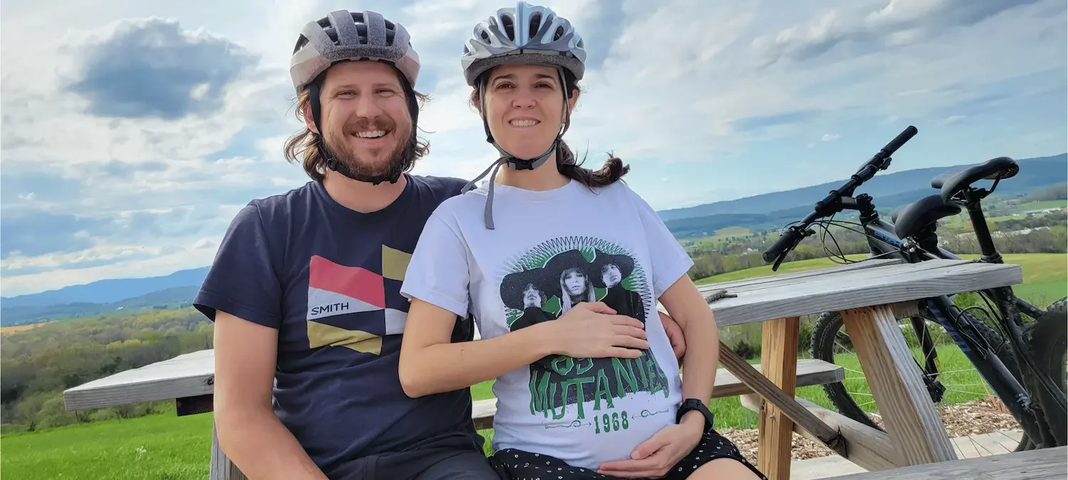 Pregnant woman and man wearing helmets smiling at camera and surrounded by mountains