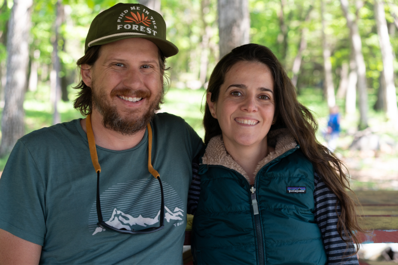 A man and woman sitting outdoors in a forested area, smiling at the camera. The man is wearing a green cap with the text 'Find Me In The Forest' and a teal t-shirt with mountain graphics. The woman is wearing a teal Patagonia vest over a striped long-sleeve shirt, with long wavy brown hair.