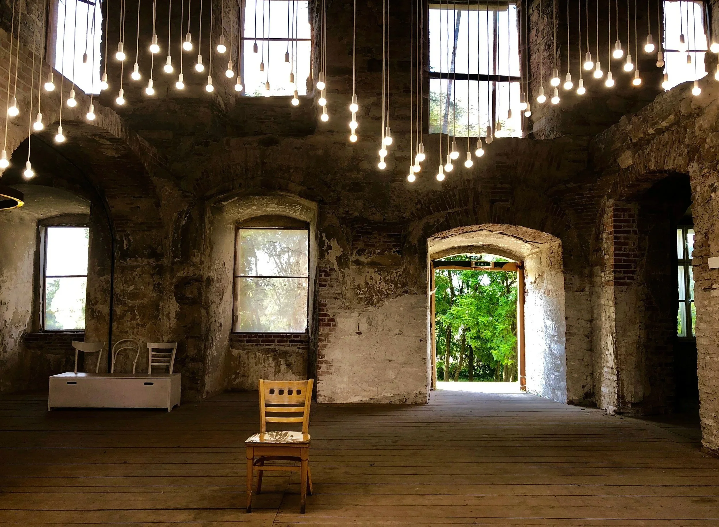 An interior view of an old, rustic brick and stone building with large windows and an open door leading outside to green trees. The ceiling has numerous hanging light bulbs, and there is a wooden chair in the center with a worn wall and floor.