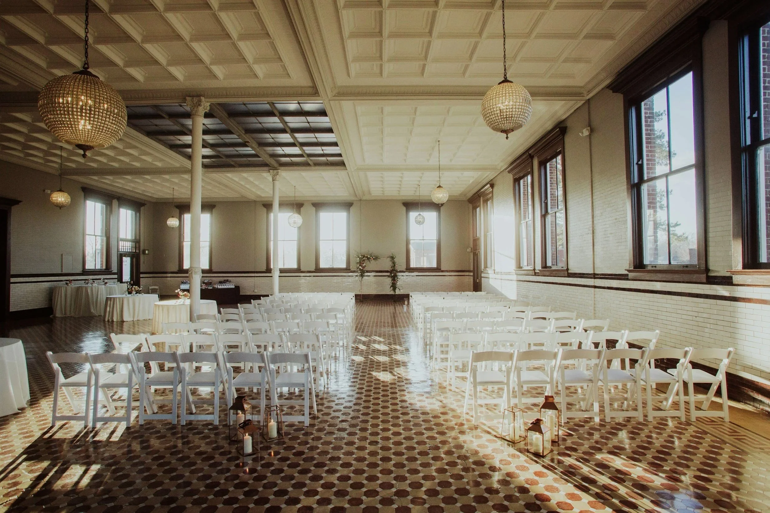 Empty wedding ceremony setup in a bright, spacious room with high ceiling, white chairs arranged in rows, large windows, and decorative hanging lights.