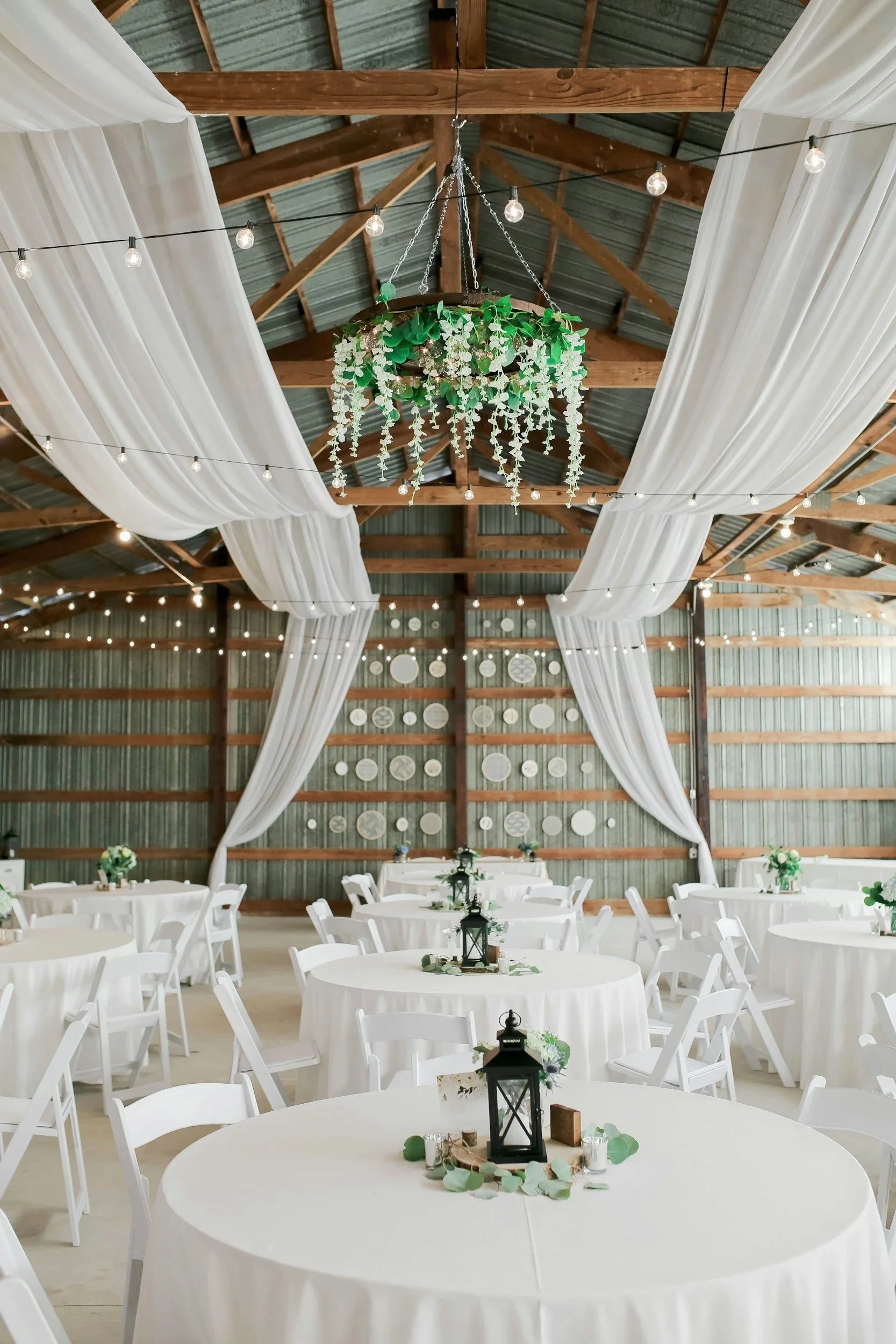 Wedding reception with round tables covered in white tablecloths, decorated with black lantern centerpieces, greenery, and candles. White chairs surround the tables. The barn interior has wooden beams, string lights, and white draped fabric, with a decorative wall of plates in the background.