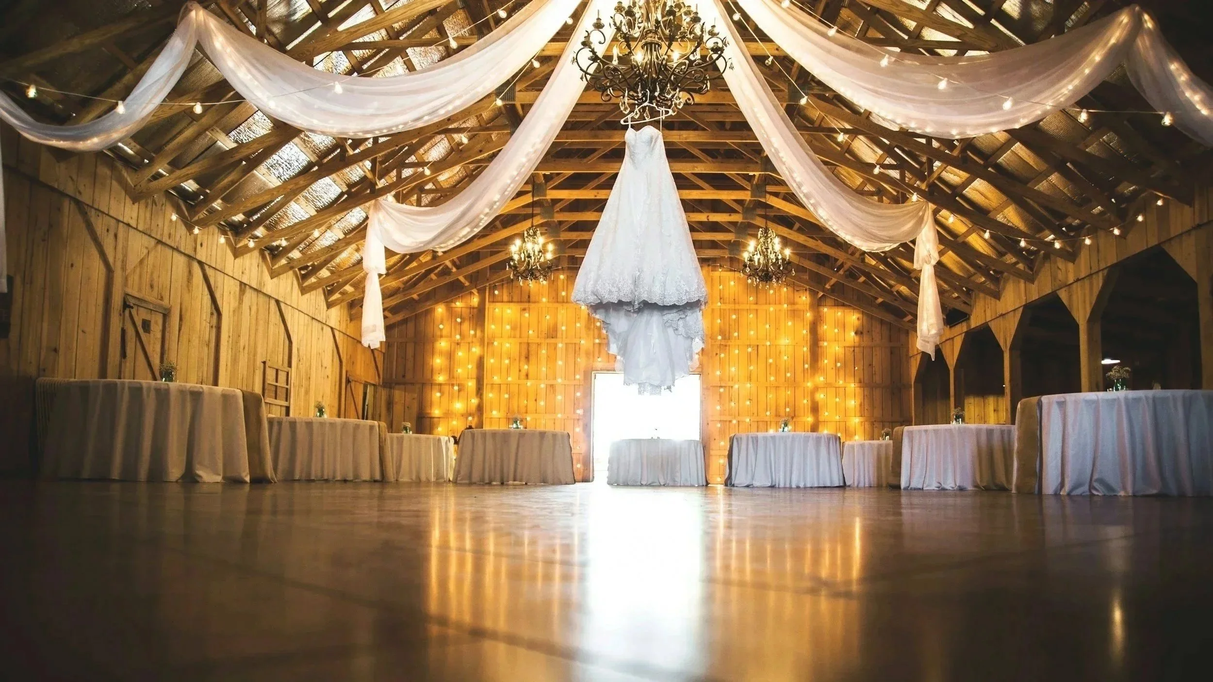 Wedding dress hanging from the ceiling in a rustic barn with string lights and chandeliers, decorated with white drapes and tables along the sides for an event.