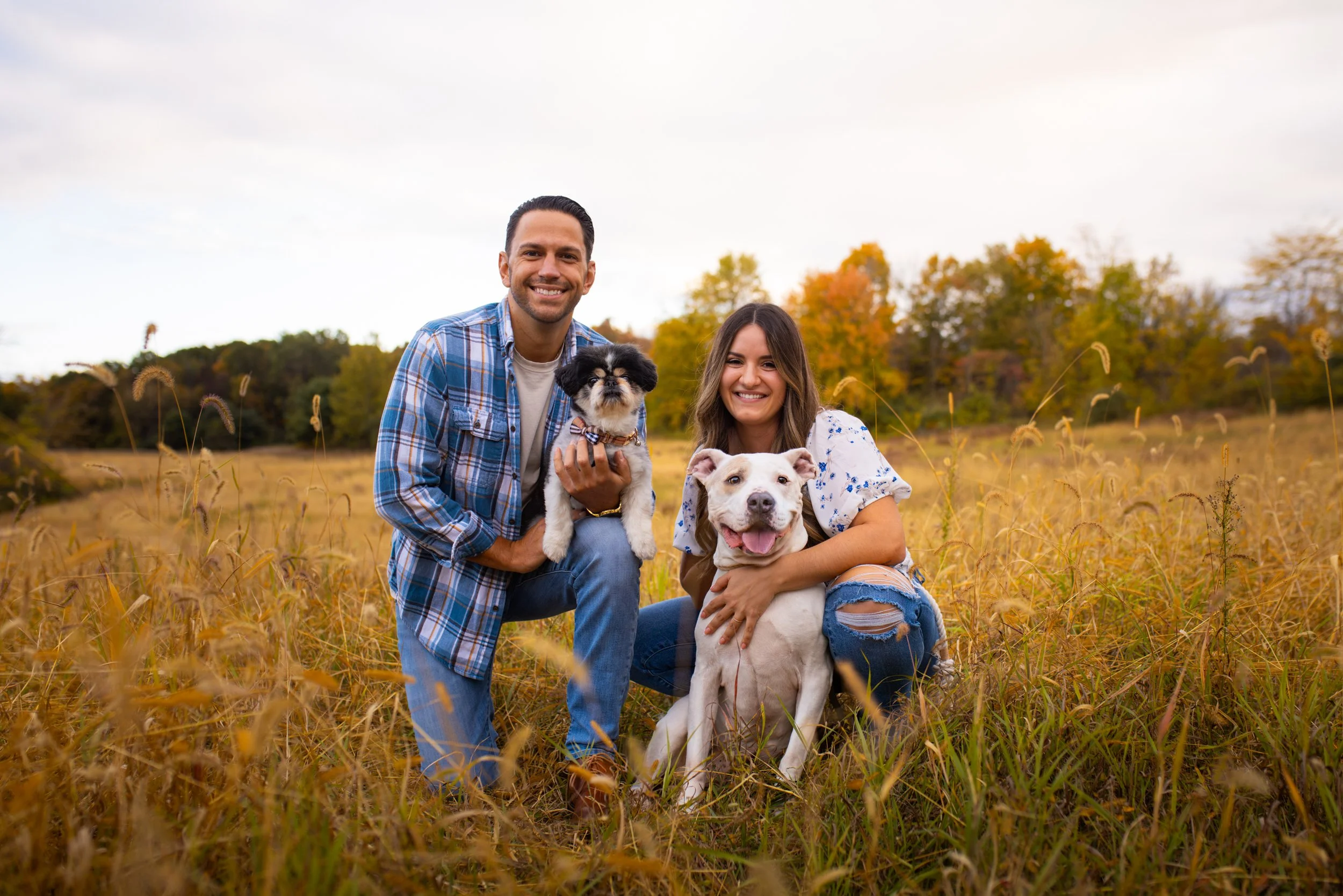 A smiling man and woman with two dogs in a grassy field during fall, with trees in the background.