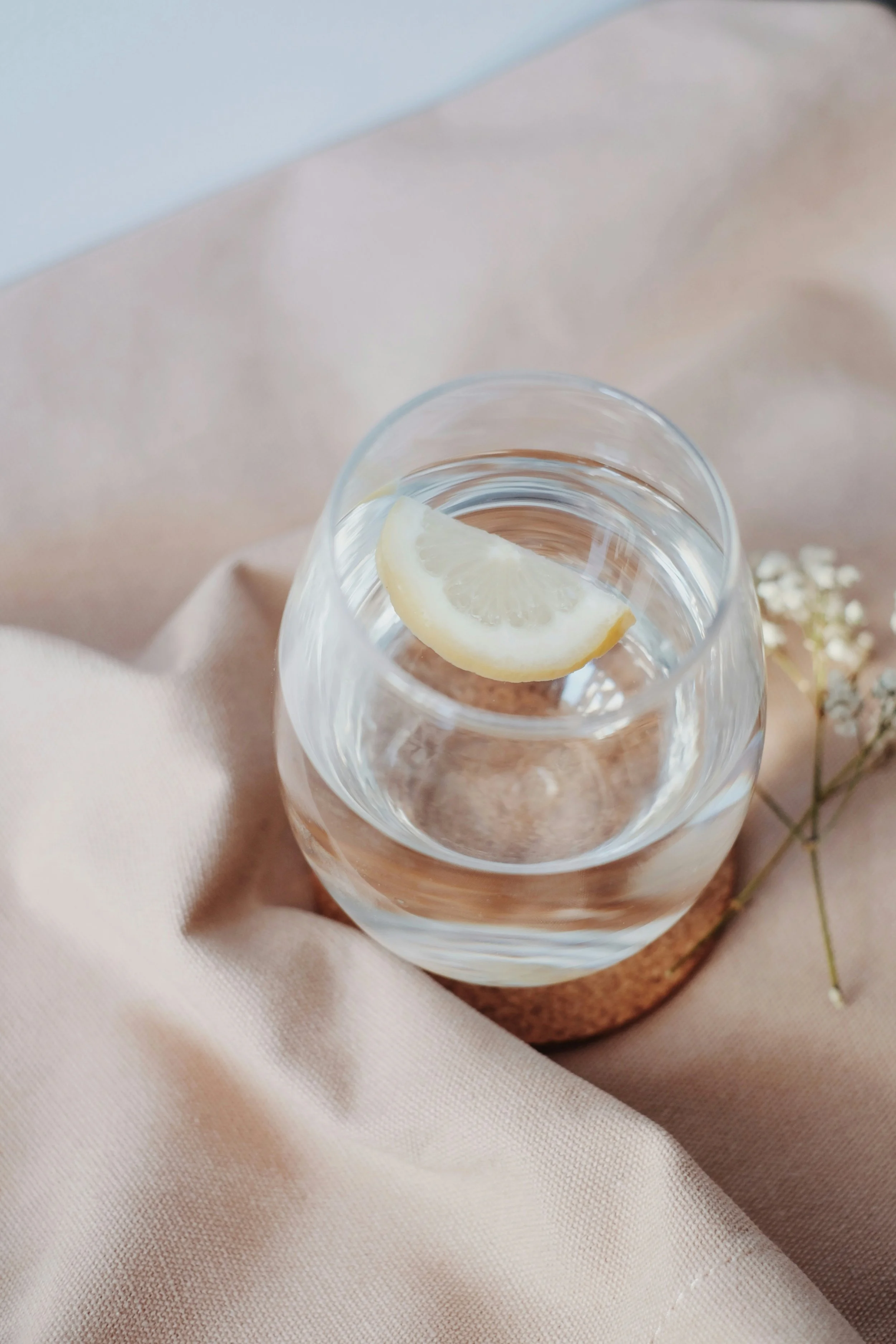 A glass of water with a lemon slice on the rim, placed on a round wooden board with a beige cloth underneath.