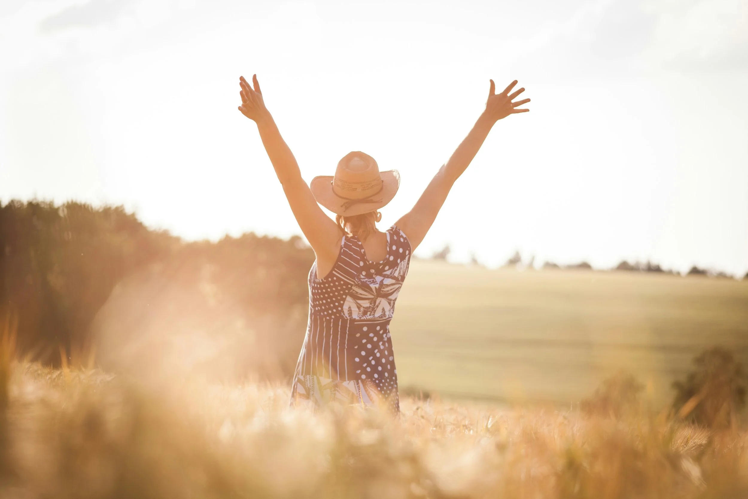 Person wearing a wide-brimmed hat and a polka-dot dress standing in a field with arms raised towards the sky during sunset.