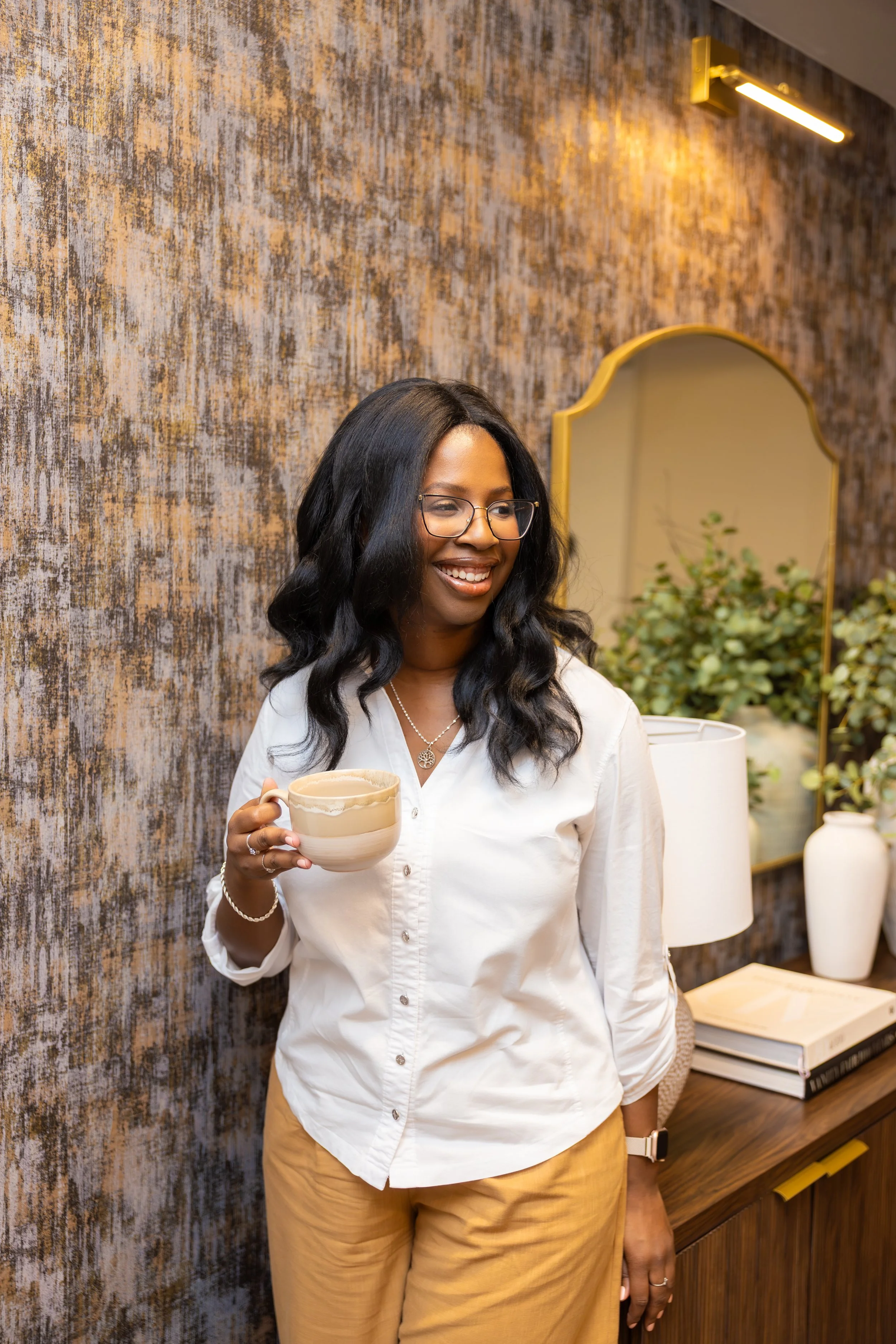 A smiling woman with glasses and long black hair holding a cup, standing in a room with textured wall and decorative mirror, wearing a white shirt and beige pants.