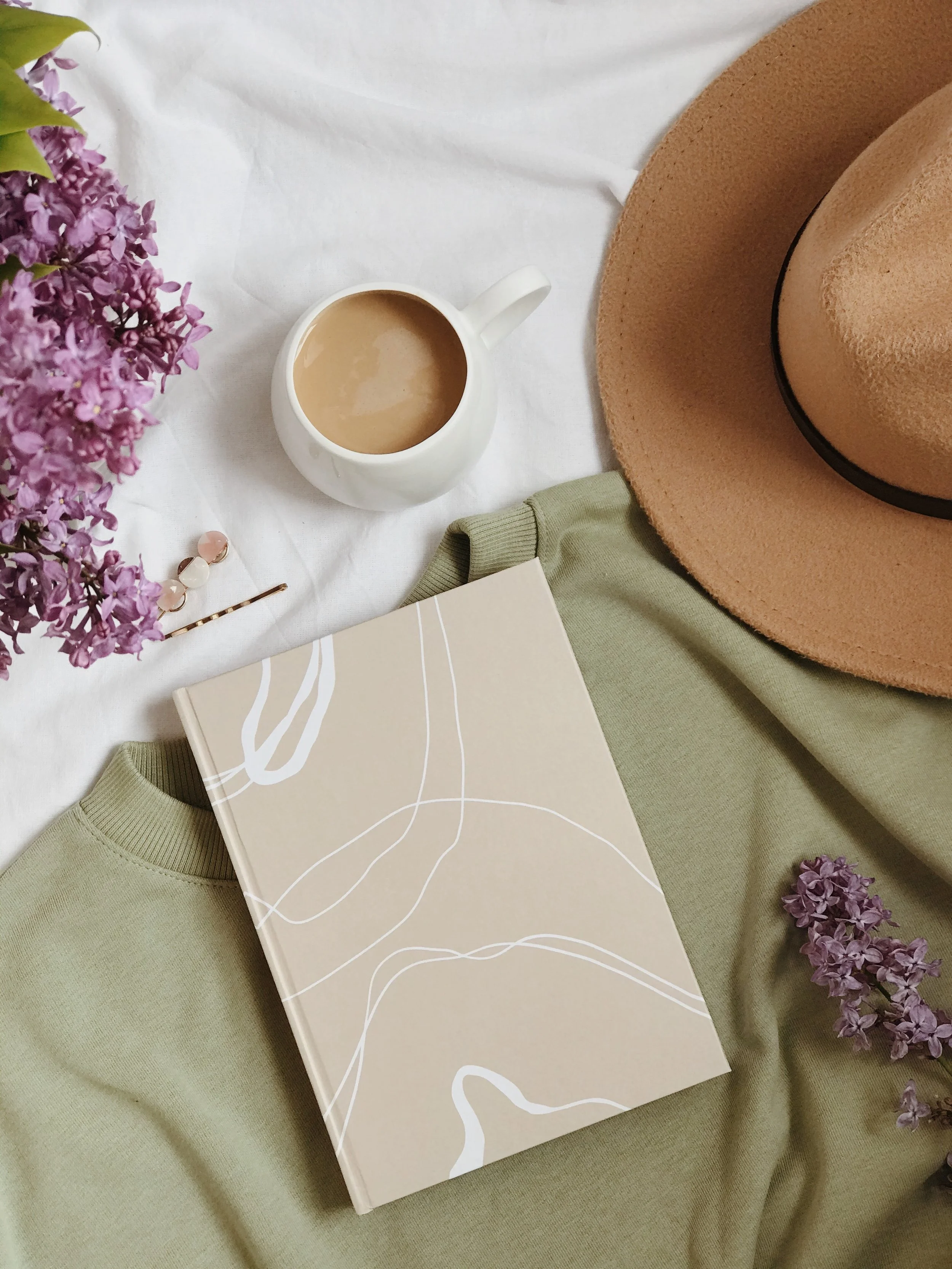 Flat lay of a beige notebook with white line art on the cover, green sweater, wide-brimmed beige hat, white coffee mug with hot beverage, purple lilac flowers, and pink and white hairpins on a white and green fabric background.