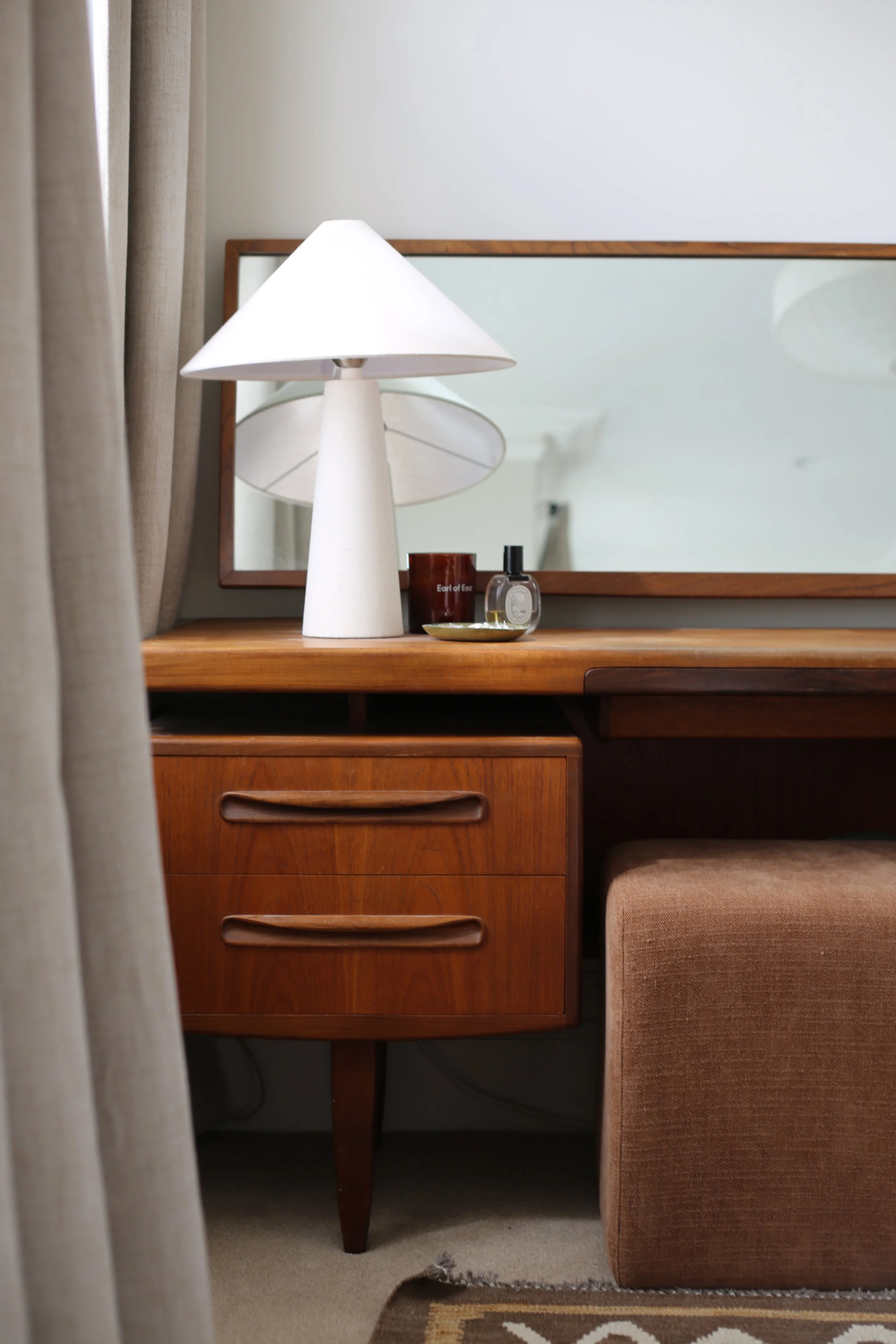 A wooden dressing table, a beige lamp, a candle and a small perfume bottle.  A curtain partially obscures the scene on the left, and the ottoman is tucked under.