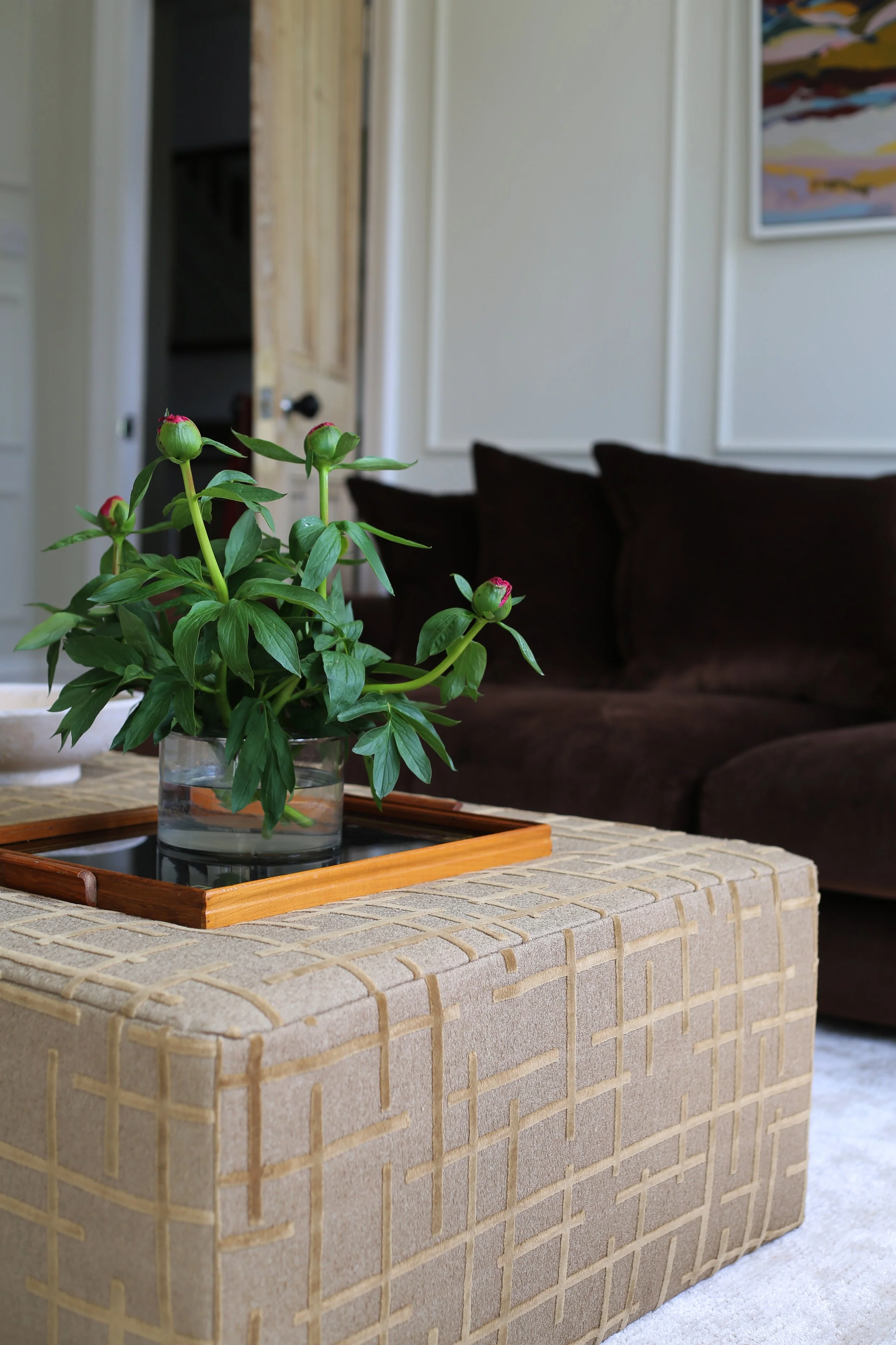 A vase of pink peonies on a tray on an upholstered ottoman, in a living room with a brown couch and a wall with artwork.