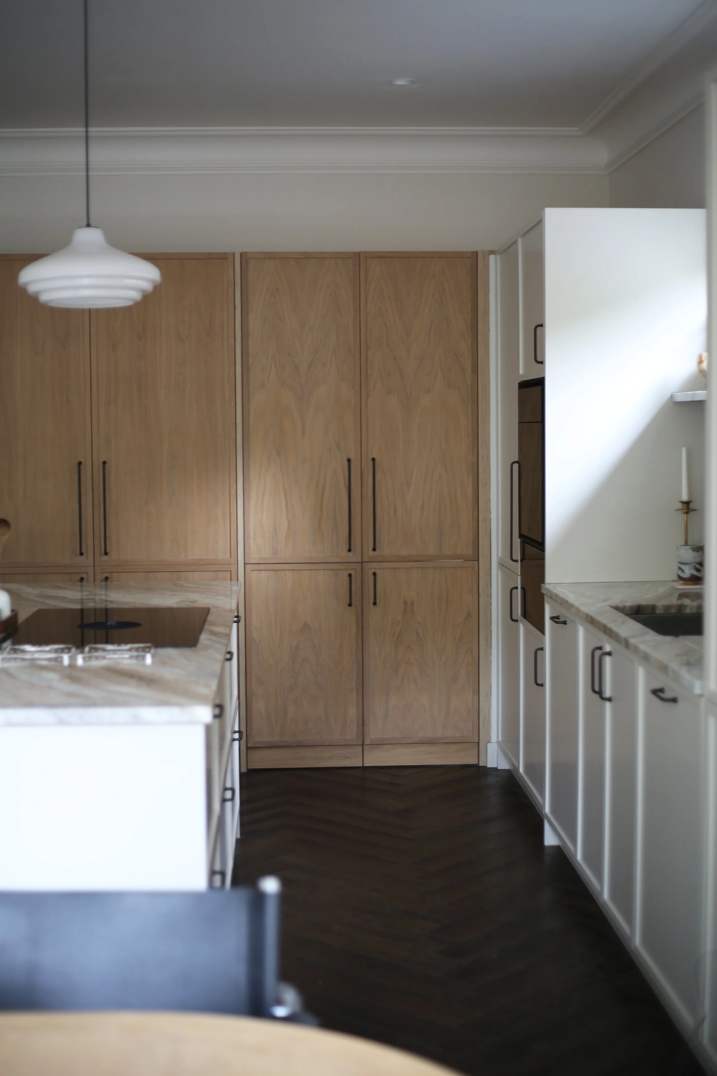 Interior view of a kitchen with wooden cabinets, a marble countertop, and a off-white wall with built-in appliances. A white hanging pendant light is visible, and a candle holder is on the counter. Dark wood flooring is present.