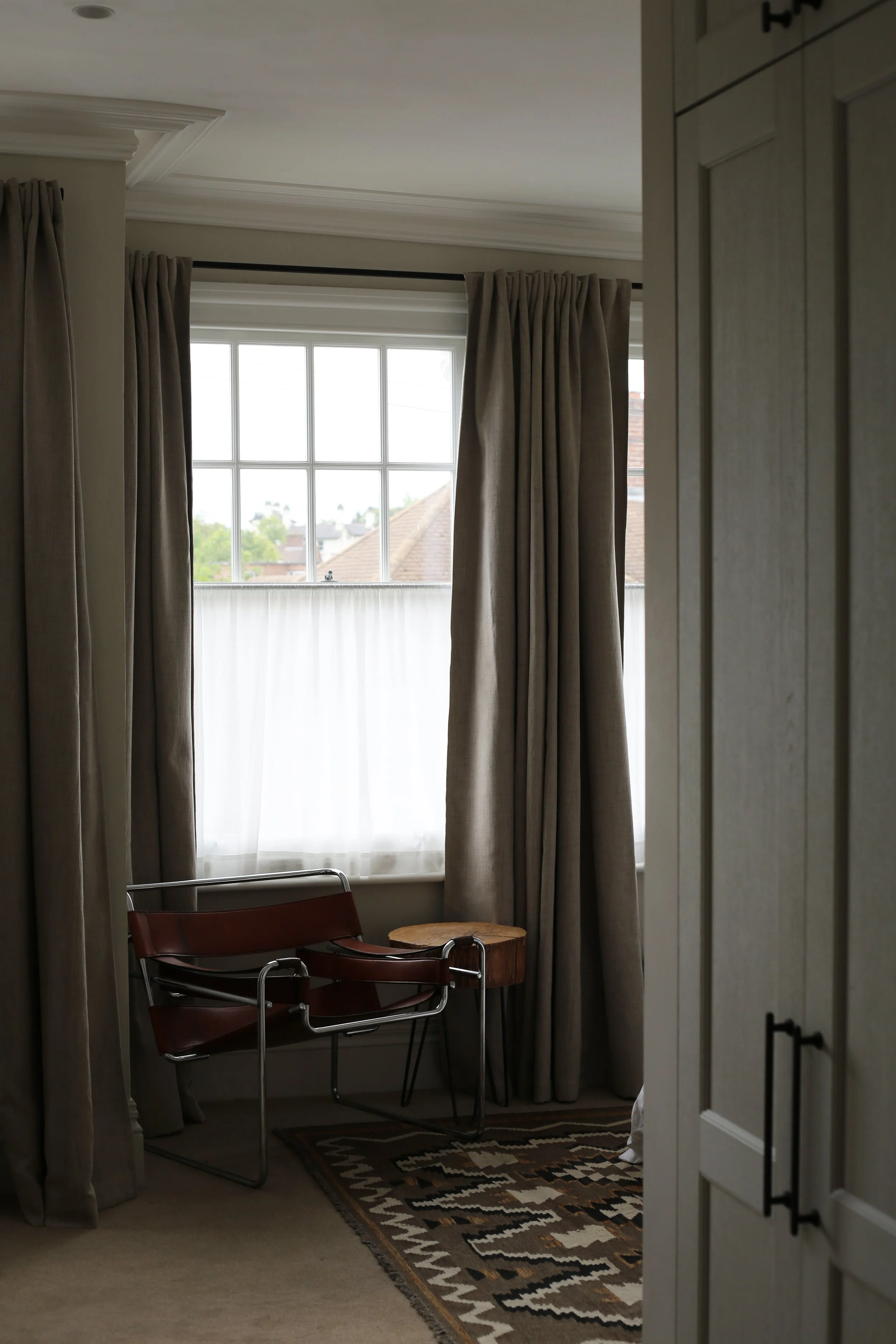 A cozy corner near a window with beige curtains, a black and chrome chair, a wooden side table, a patterned rug, and a closet door.
