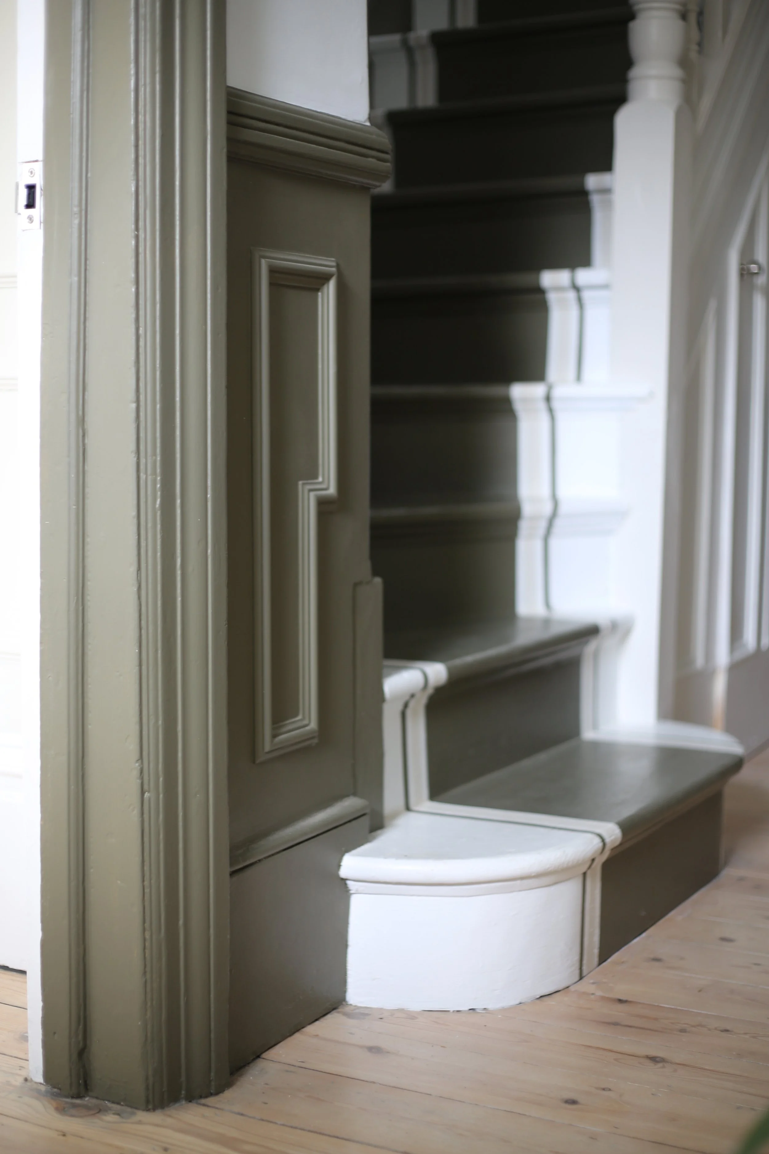 A vintage staircase with painted dark stripe steps and a white curved base, partially hidden behind a door frame on the left, set on a wooden floor.