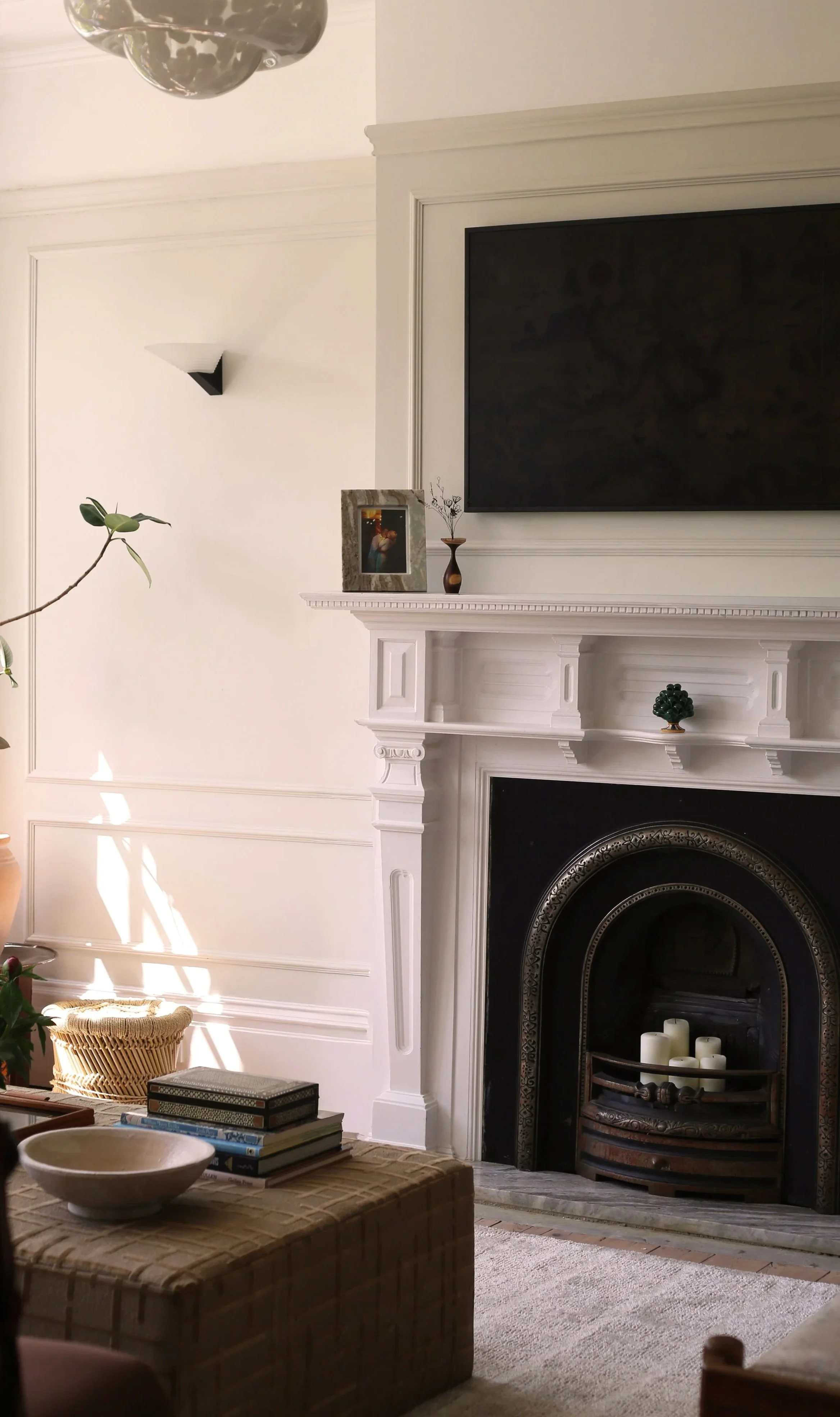 Living room with neutral walls, a fireplace with black and white border, a mounted flat-screen TV, and decorative items on the mantel and upholstered ottoman .